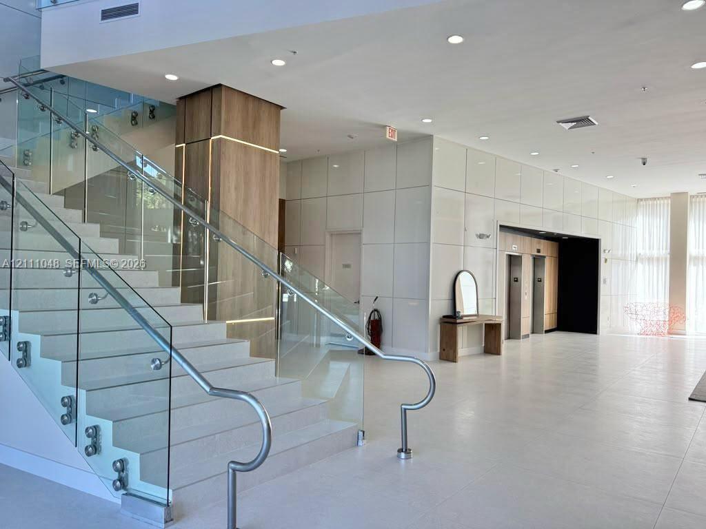 Lobby area featuring a wide staircase with glass railings and metal handrails, a central pillar with integrated accent lighting and wood-finish paneling, large-format tiled flooring, and high-gloss wall panels Lobby area featuring a wide staircase with glass railings and metal handrails, a central pillar with integrated accent lighting and wood-finish paneling, large-format tiled flooring, and high-gloss wall panels