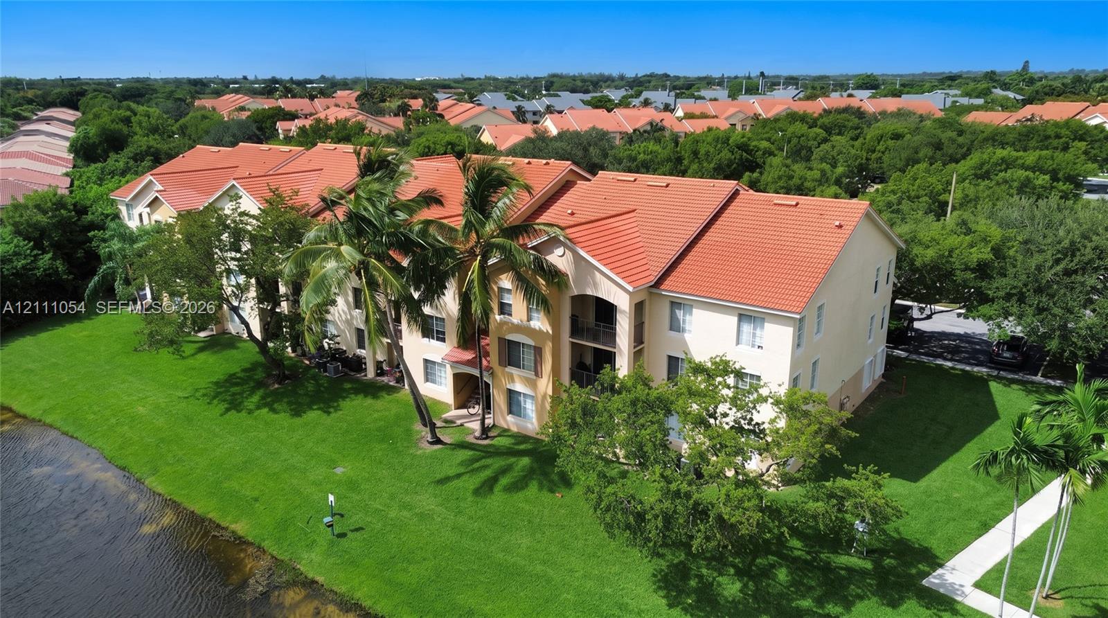 Multi-story building with terracotta-style roof tiles, stucco exterior, private balconies, and waterfront positioning surrounded by mature landscaping