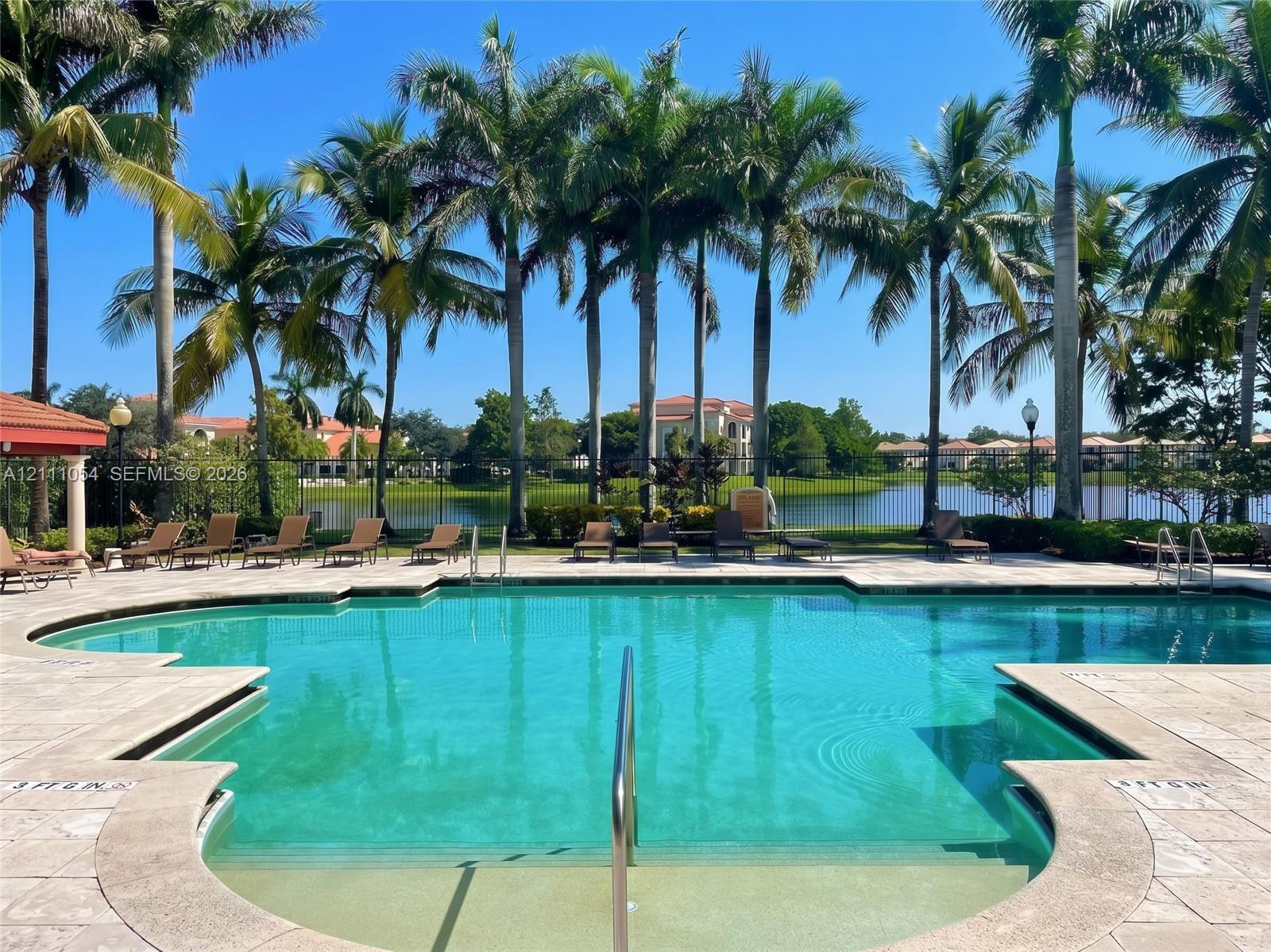 Swimming pool with stone paver decking, metal perimeter fencing, and surrounding mature palm trees overlooking a lake