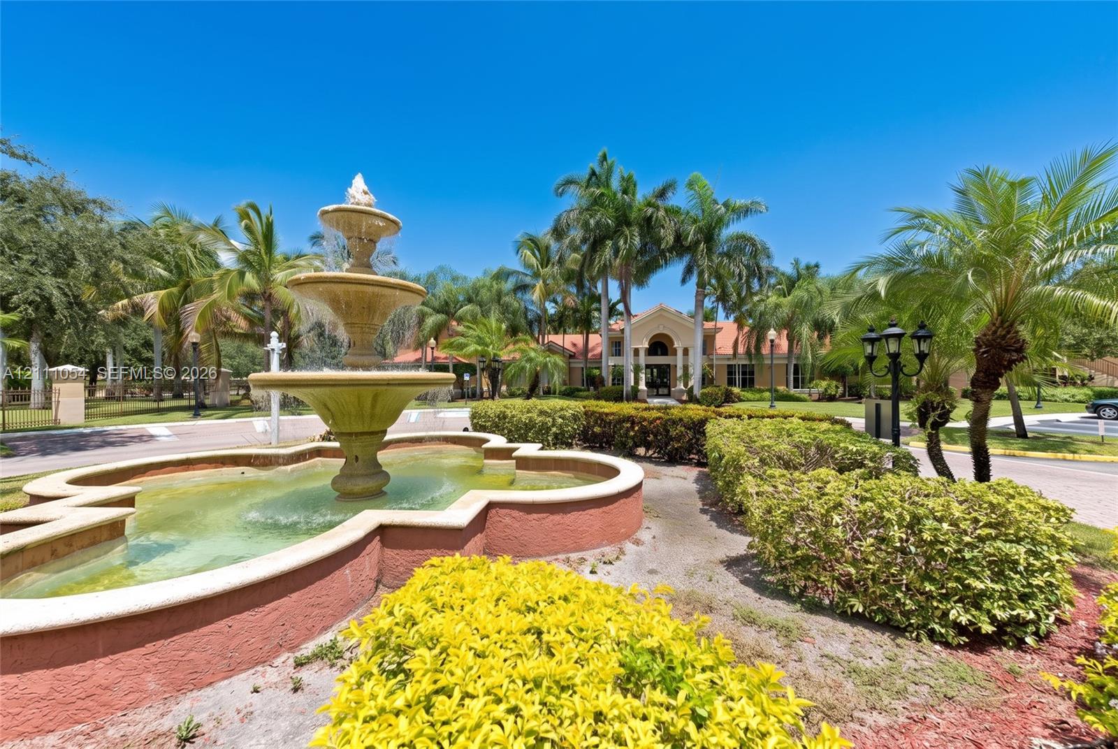 Tiered stone water fountain featuring a decorative basin, surrounded by professional landscaping, manicured hedges, and mature palm trees
