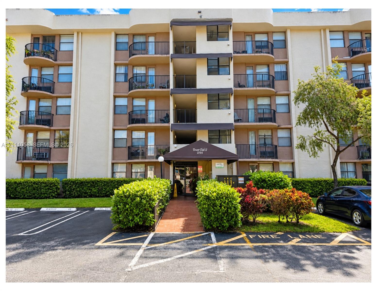 Multi-story residential building exterior featuring curved private balconies, metal railings, and a covered entryway with a brick-paved walkway Multi-story residential building exterior featuring curved private balconies, metal railings, and a covered entryway with a brick-paved walkway