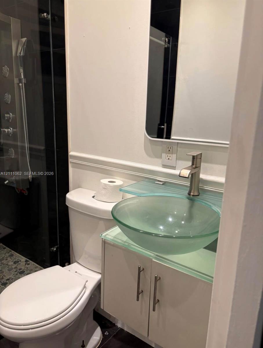 Frosted glass vessel sink with a glass countertop, chrome-finish faucet, white cabinetry with metal hardware, and a glass-enclosed shower featuring a multi-head fixture