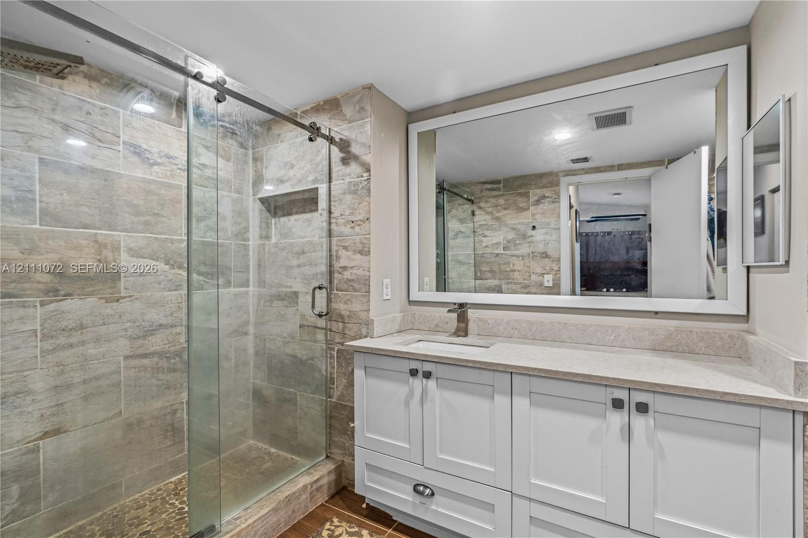 Walk-in shower with stone-tile surround, glass sliding door, and integrated shower niche, paired with a white Shaker-style vanity featuring a stone countertop and large framed wall mirror