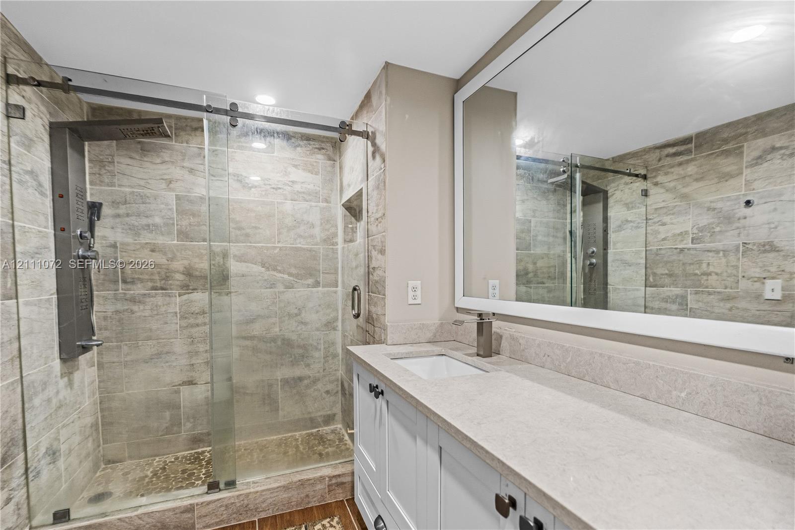 Bathroom featuring a walk-in shower with sliding glass doors, stone-finish wall tile, a built-in shower panel, white cabinetry with a stone-finish countertop, and a large framed vanity mirror