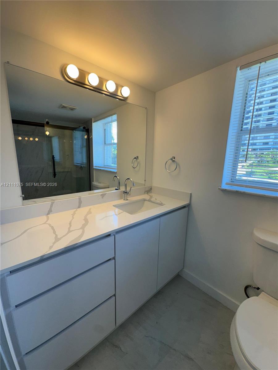 Bathroom featuring a vanity with a white-veined stone countertop, an integrated rectangular sink, wall-to-wall mirror, four-bulb vanity lighting, and a walk-in shower with glass enclosure