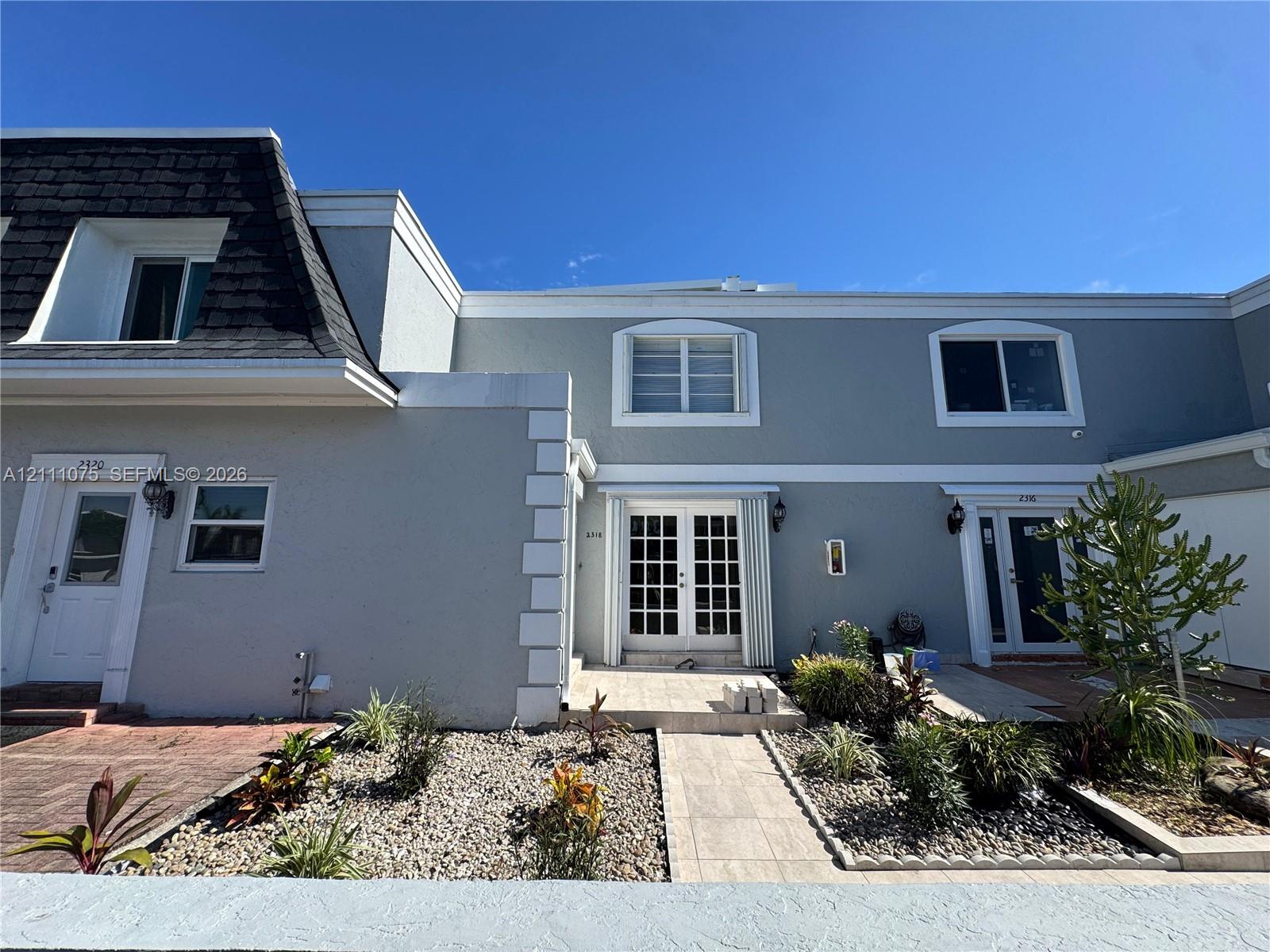 Exterior facade featuring a combination of stucco and shingled roofline, paired glass entry doors, arched window detailing, and xeriscaped landscaping with stone hardscaping