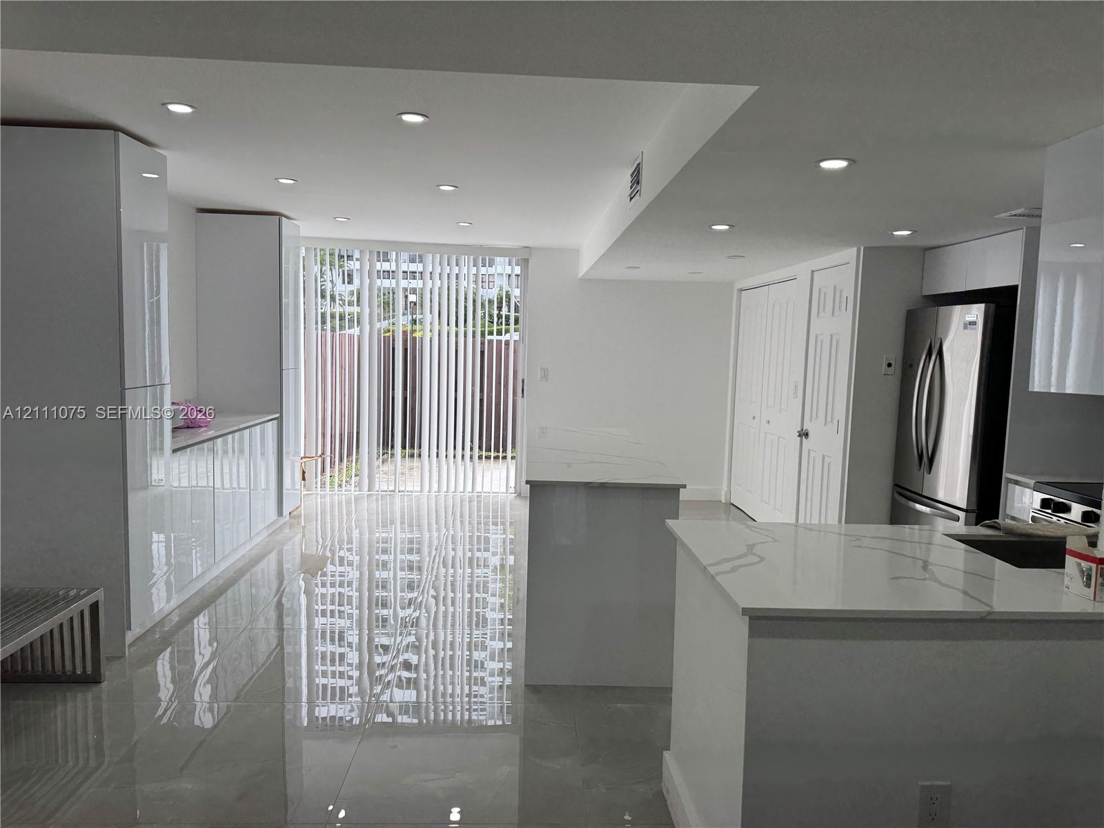 Modern kitchen featuring high-gloss cabinetry, stone-surface countertops, polished tile flooring, recessed ceiling lighting, and a stainless steel refrigerator Modern kitchen featuring high-gloss cabinetry, stone-surface countertops, polished tile flooring, recessed ceiling lighting, and a stainless steel refrigerator