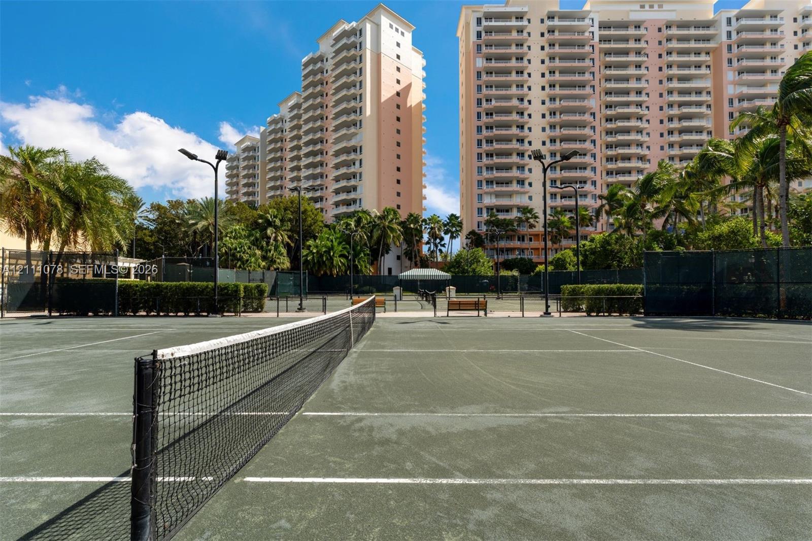 Tennis court featuring a synthetic surface, perimeter fencing, overhead lighting, and benches