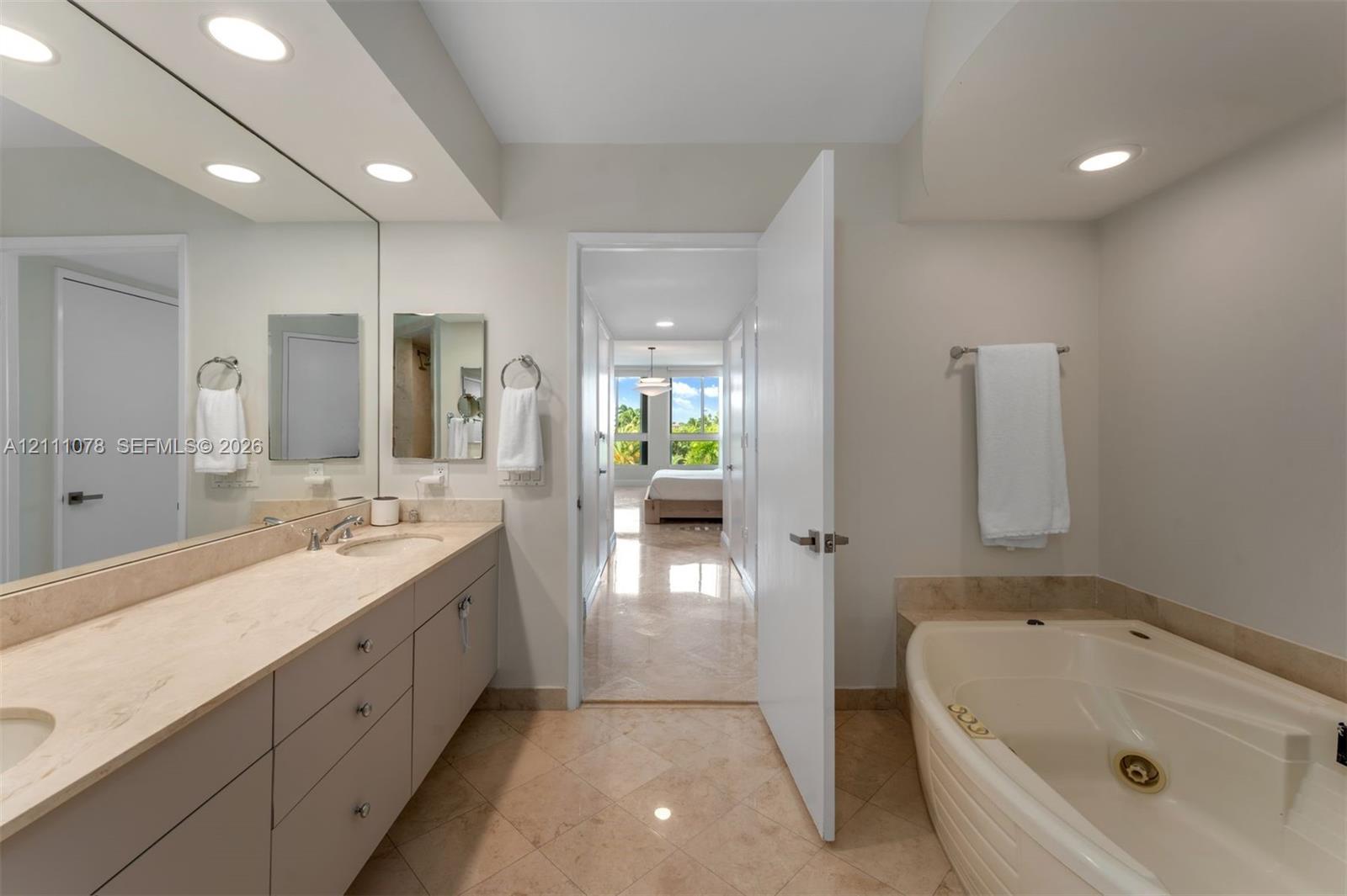 Bathroom featuring dual-sink vanity with stone countertop, large wall-mounted mirror, recessed lighting, tile flooring, and jetted tub with stone tile surround