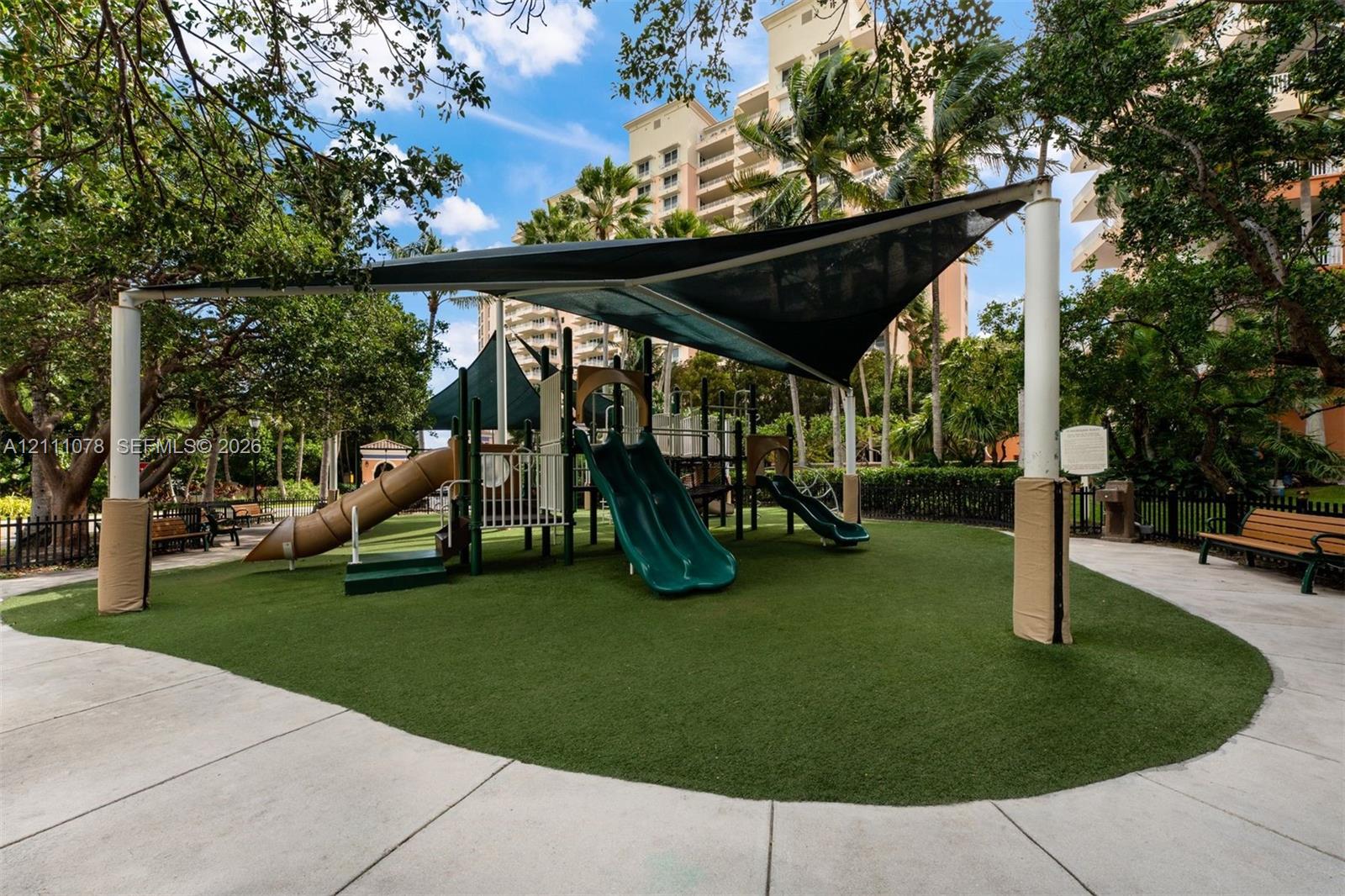 Recreational area featuring a playground structure with multiple slides, synthetic turf surfacing, a canopy shade sail, and surrounding concrete pathways
