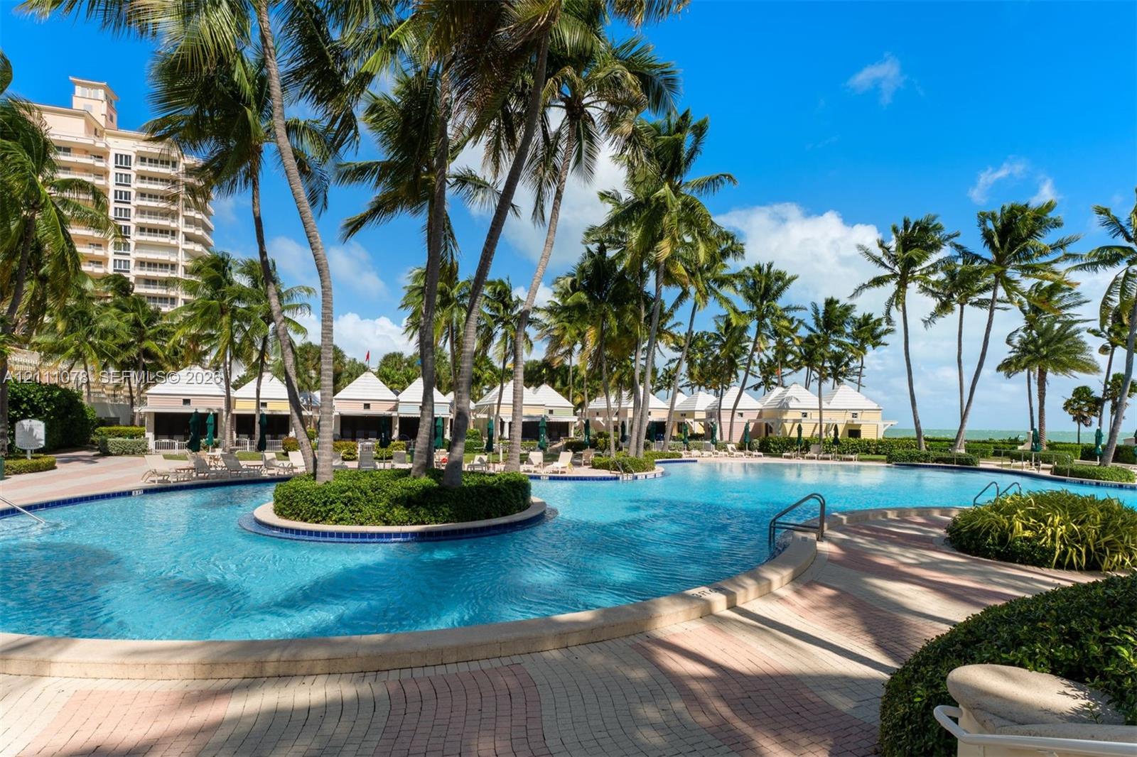 Resort-style swimming pool featuring a central island with mature palm trees, multi-tone paver decking, and cabanas