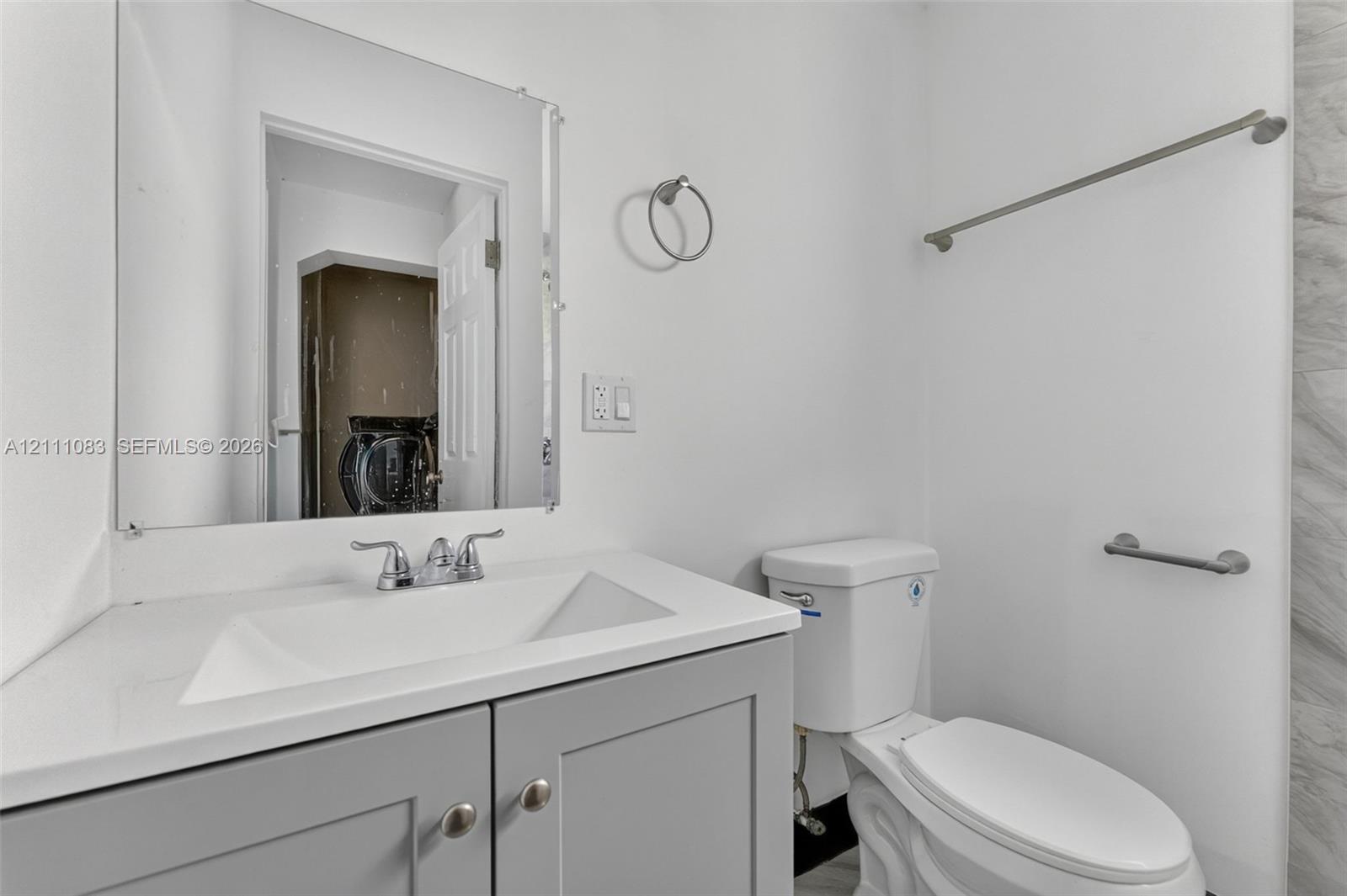Grey shaker-style vanity with a solid surface countertop, integrated sink basin, polished chrome faucet, rectangular frameless mirror, and brushed nickel hardware