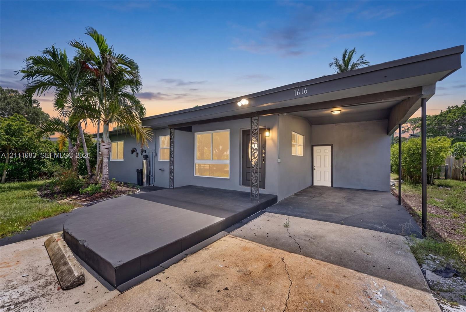 Single-story exterior featuring a grey stucco facade, covered carport, raised concrete entrance platform, and decorative metal support columns