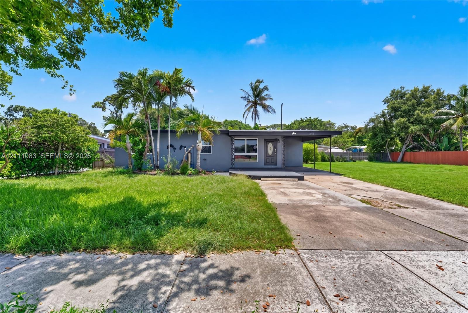 Single-story exterior featuring a flat roofline, attached covered carport, concrete driveway, and established palm trees