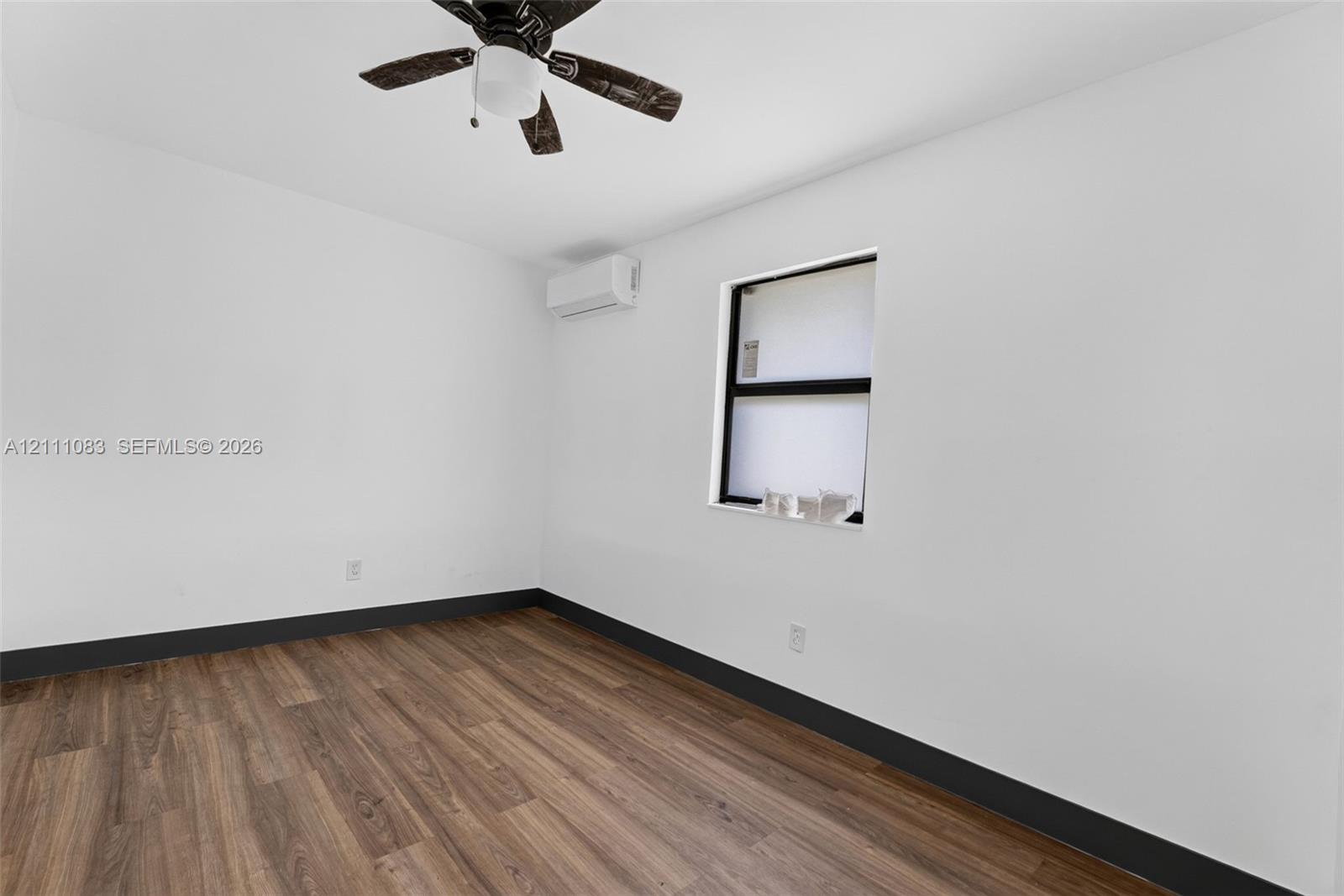 Room with wood-finish flooring, ceiling fan with light fixture, window with black frame, and wall-mounted climate control unit