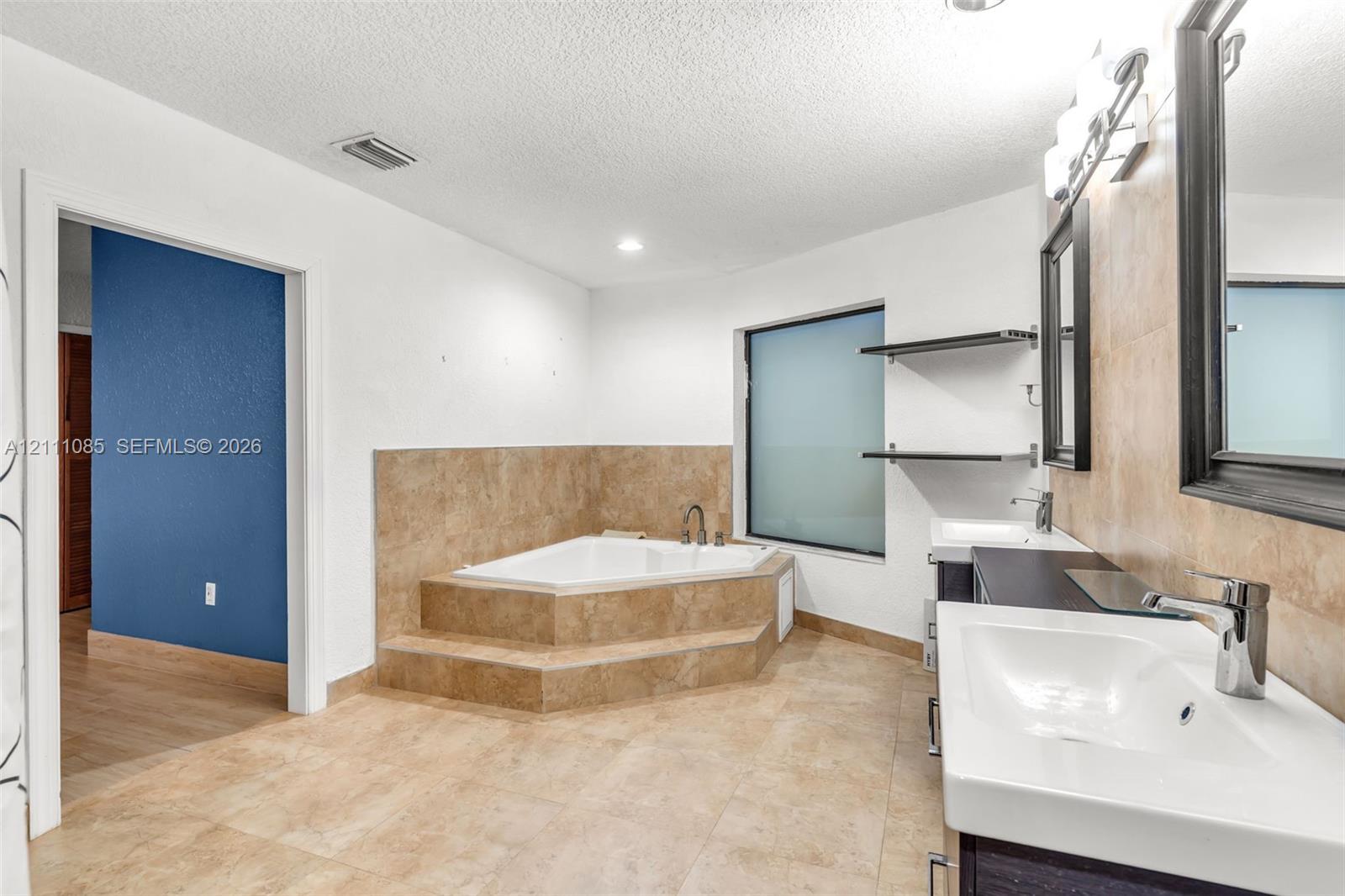 Bathroom featuring a corner soaking tub with tile surround, dual-sink vanity with dark cabinetry, wall-mounted shelving, and tile flooring