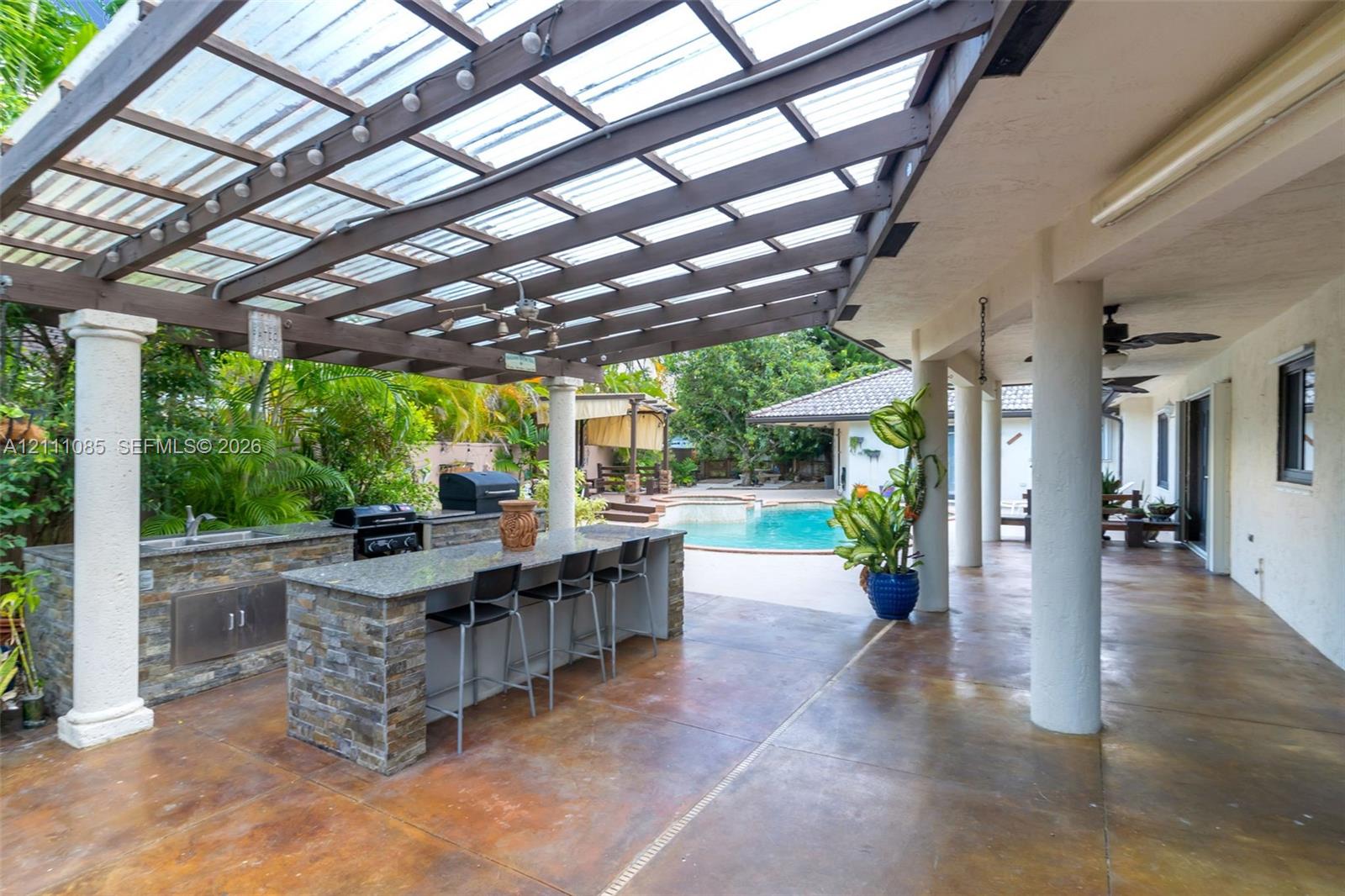 Covered patio featuring an outdoor kitchen with stone veneer base, countertop bar seating, a slatted timber pergola, and a concrete pool deck