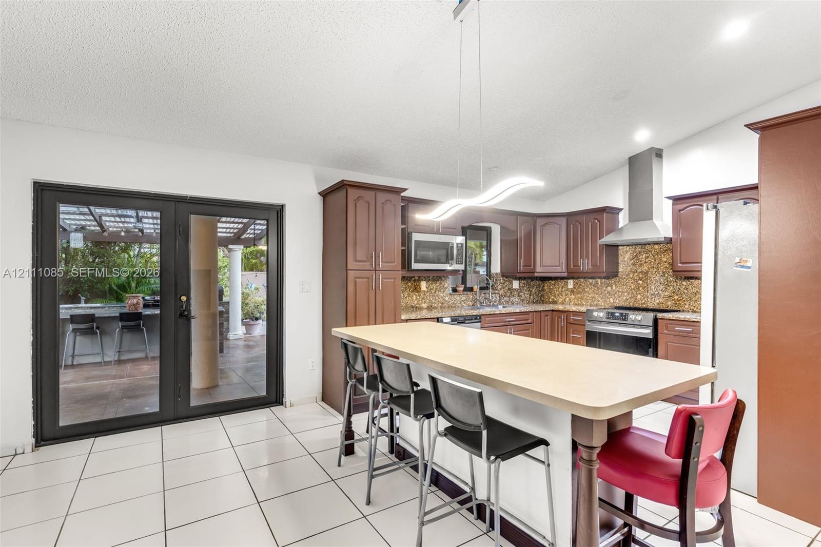 Kitchen featuring dark cabinetry, stone-finish backsplash, stainless steel range hood, central island with seating counter, and dual glass-panel doors