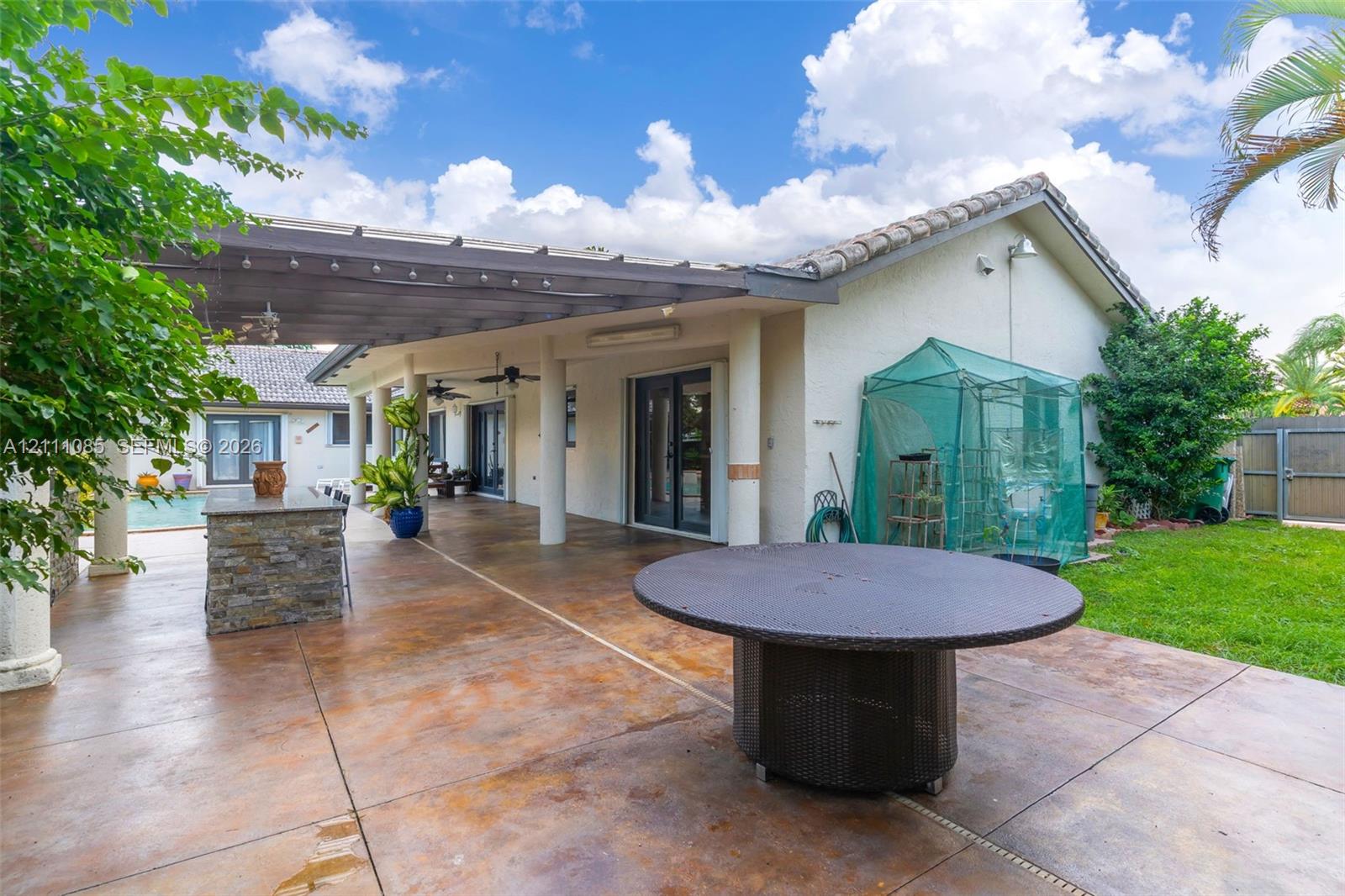 Covered concrete patio featuring a stone-clad outdoor kitchen, structural columns, ceiling fans, and built-in lighting