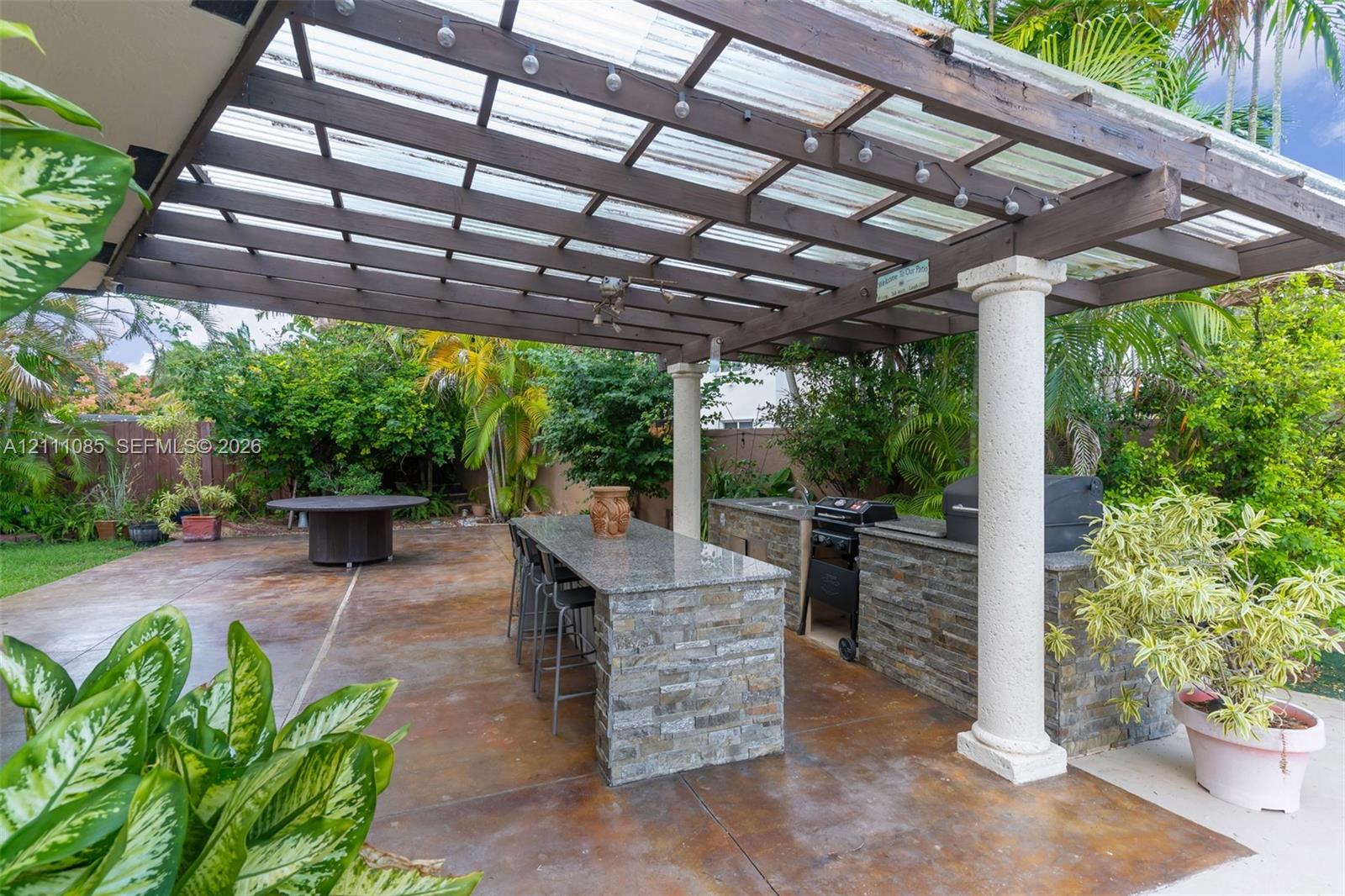 Outdoor covered patio featuring a timber-frame pergola with a corrugated roof, stone-clad outdoor kitchen island with a stone countertop, and decorative structural columns