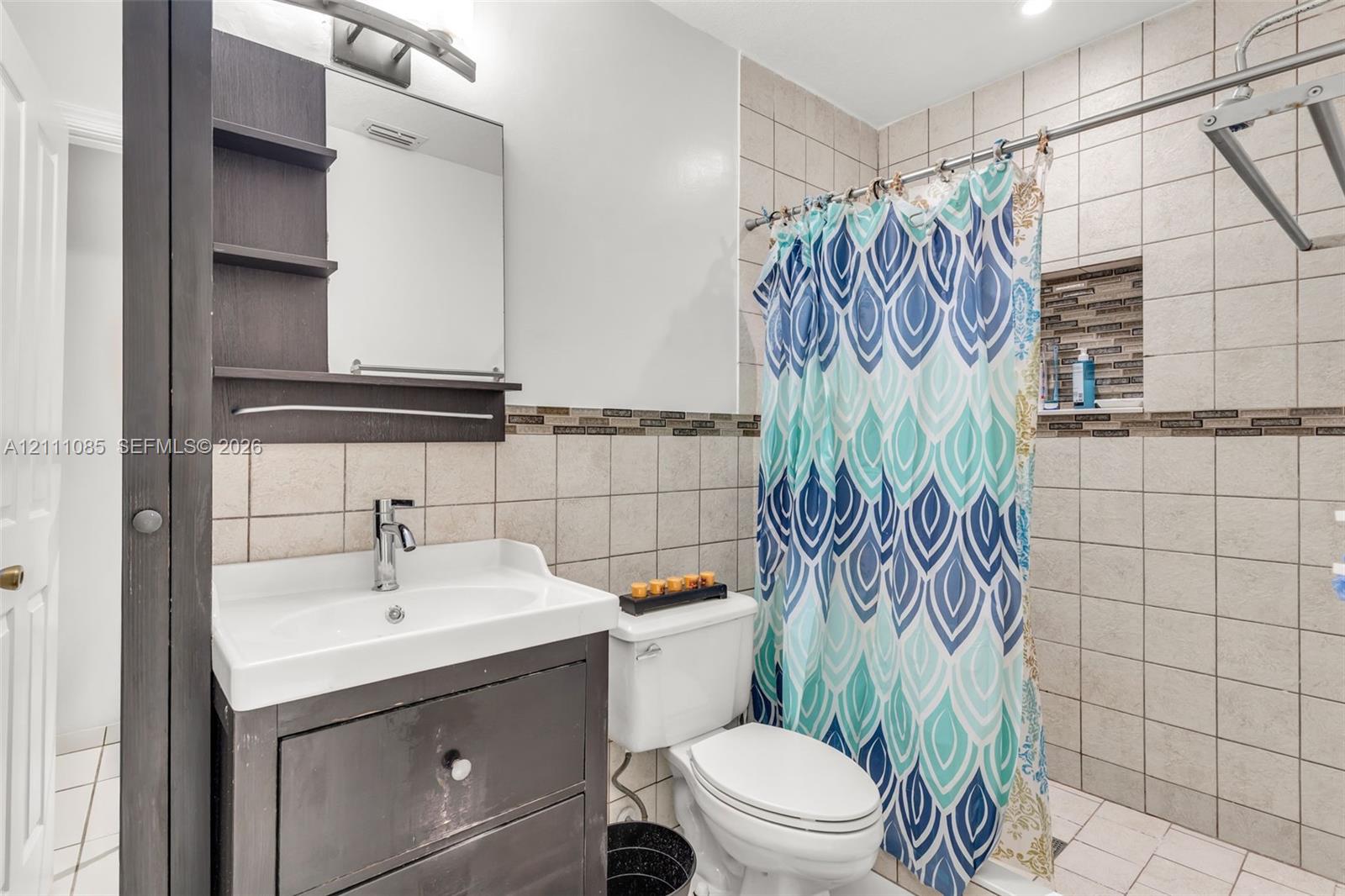 Dark-finished vanity with an integrated mirror and shelving, square wall tiling, decorative mosaic tile accents, recessed shower niche, and a standard toilet