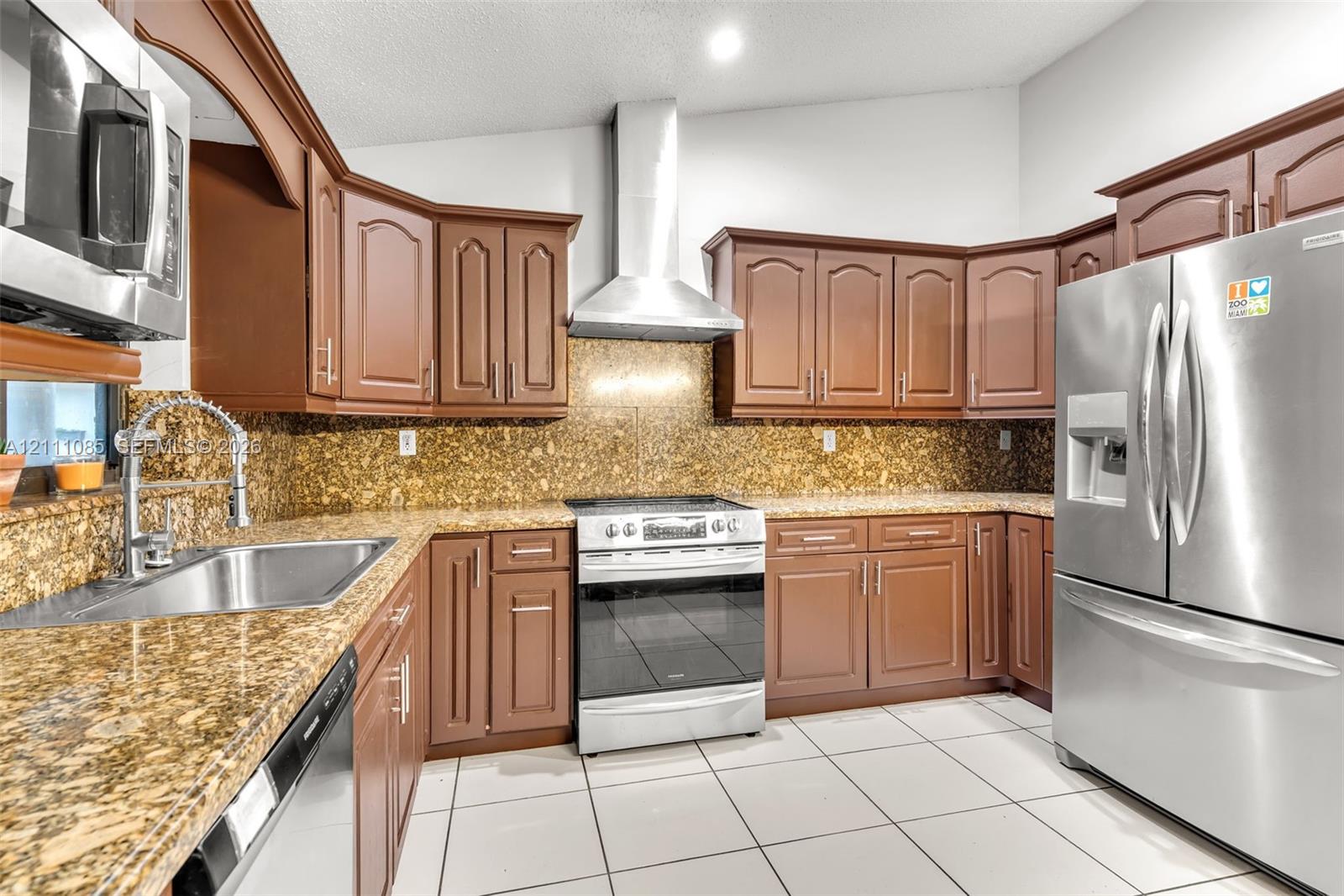 Kitchen featuring stained wood cabinetry, stone-finish countertops, stainless steel appliances, a chimney-style range hood, and tile flooring