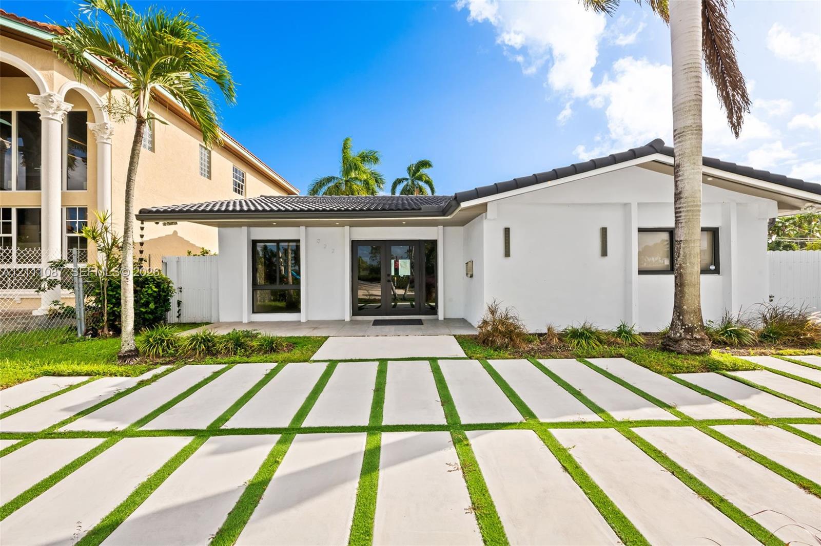 Single-story exterior featuring white stucco siding, a tiled roof, dual-entry glass doors, and an expansive concrete paver driveway with integrated grass turf