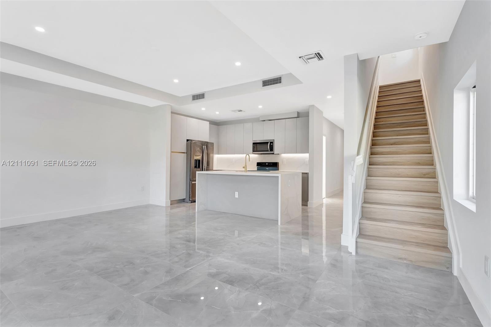 Kitchen featuring light-toned cabinetry, quartz waterfall-edge island, and stainless-steel appliances, complemented by large-format tile flooring and wood-finish staircase