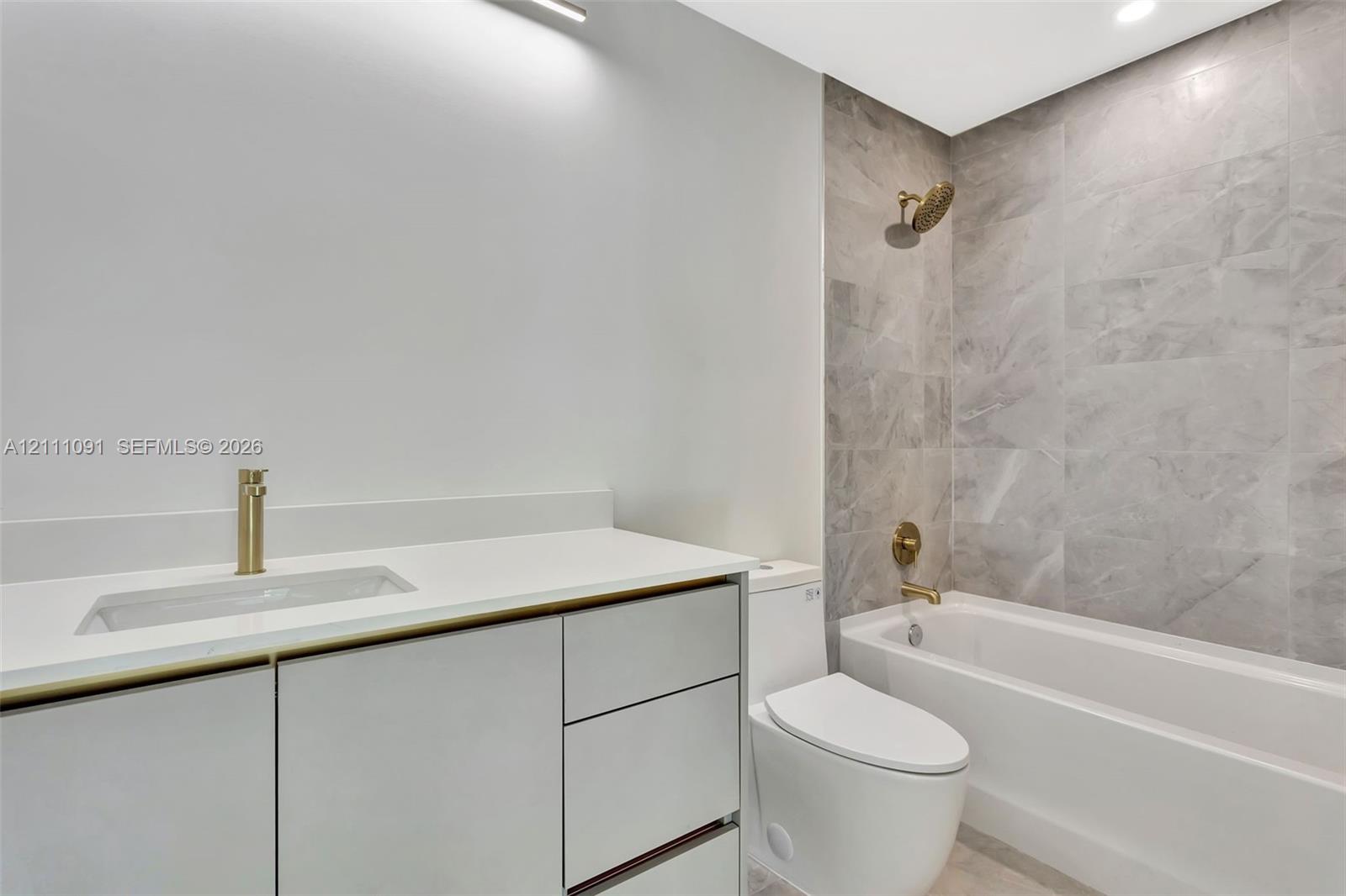 Bathroom featuring a vanity with quartz countertop, light-toned cabinetry, brass-finish fixtures, and a tub enclosure with large-format tile surround