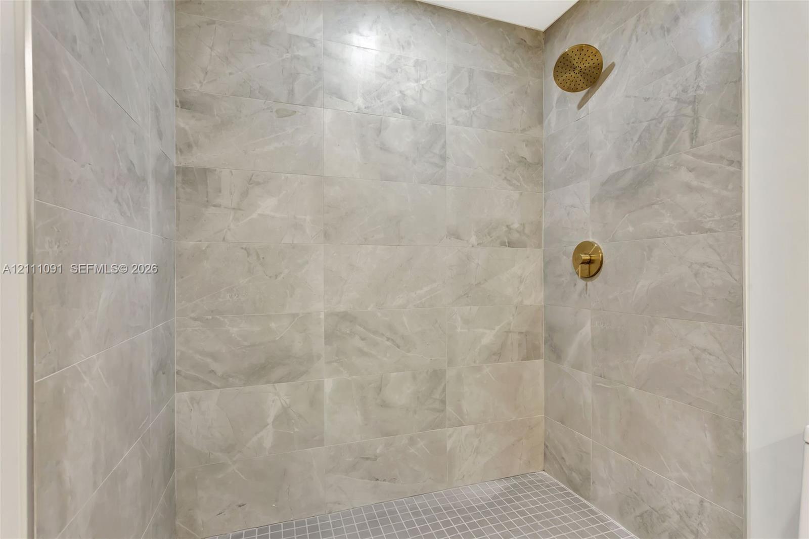 Walk-in shower featuring large-format stone-finish wall tile, a mosaic tile floor, and brushed-gold hardware including a rainfall showerhead and wall-mounted control