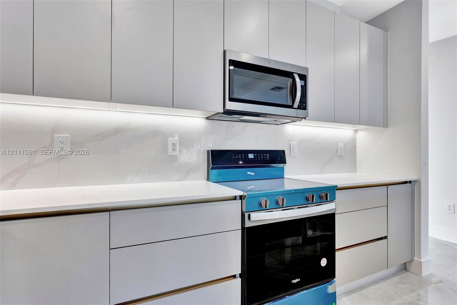 Sleek light-gray flat-panel cabinetry, white stone-finish slab backsplash, integrated under-cabinet lighting, solid-surface countertops, and stainless steel appliances