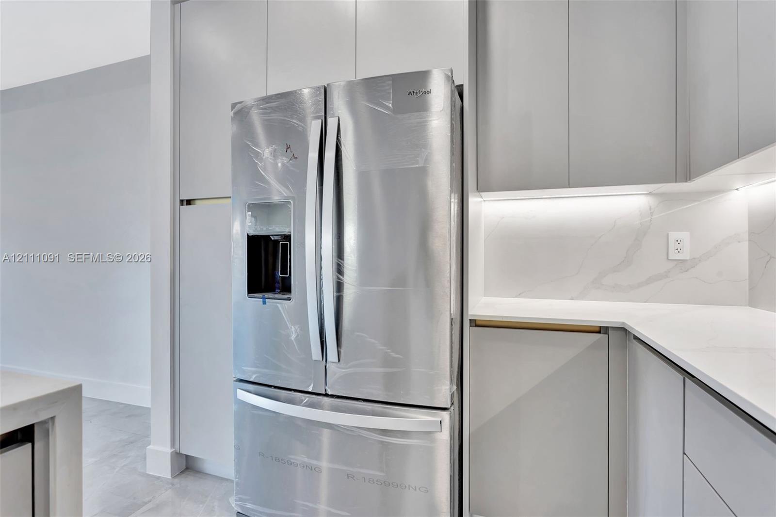 Stainless steel French door refrigerator set within floor-to-ceiling cabinetry, featuring light-toned slab-panel doors, stone-composite countertops, marble-patterned backsplash, and under-cabinet integrated lighting