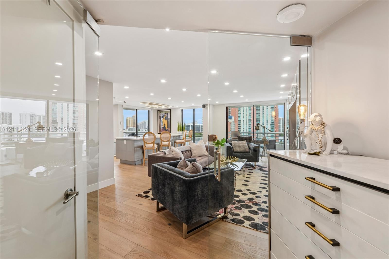 Frameless glass pivot door, light-toned wood-finish flooring, recessed ceiling lighting, floor-to-ceiling windows, and built-in cabinetry with matte finish hardware