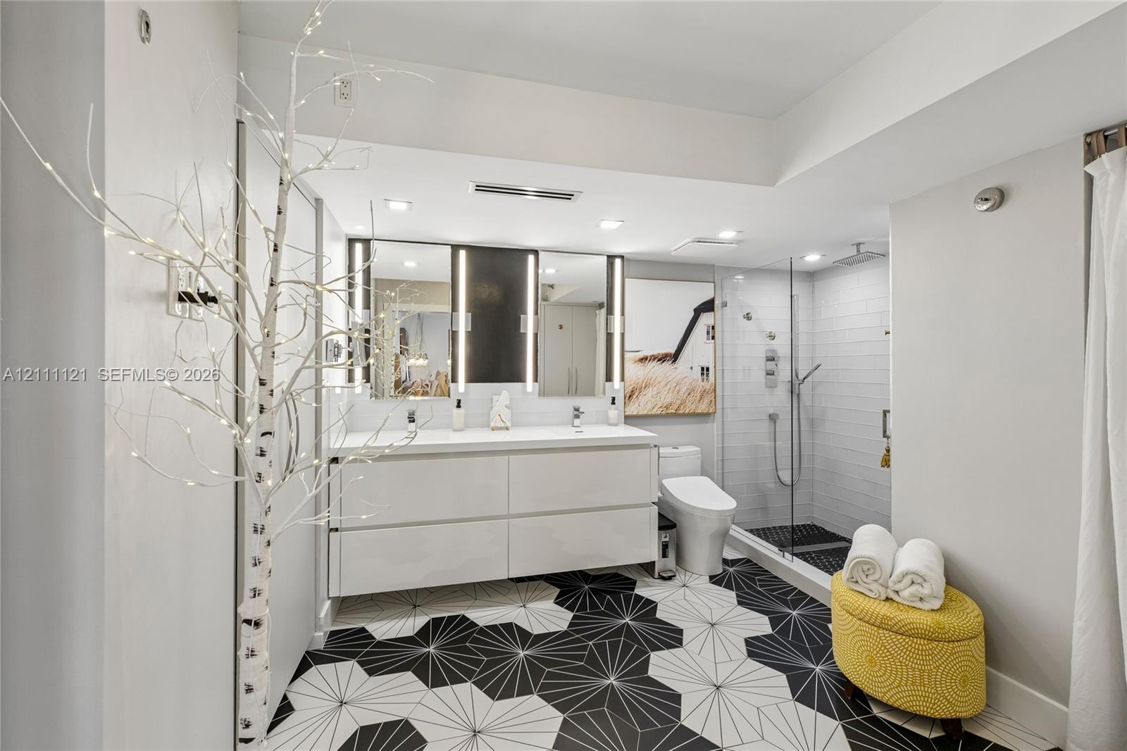 Floating double vanity with integrated sinks, geometric patterned floor tiling, frameless glass-enclosed shower with wall-mounted fixtures, and dual vertical-light vanity mirrors
