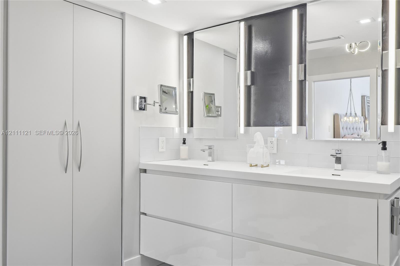 Dual-sink vanity featuring a white countertop, glossy white cabinetry, integrated LED mirror lighting, and a full-height built-in storage cabinet