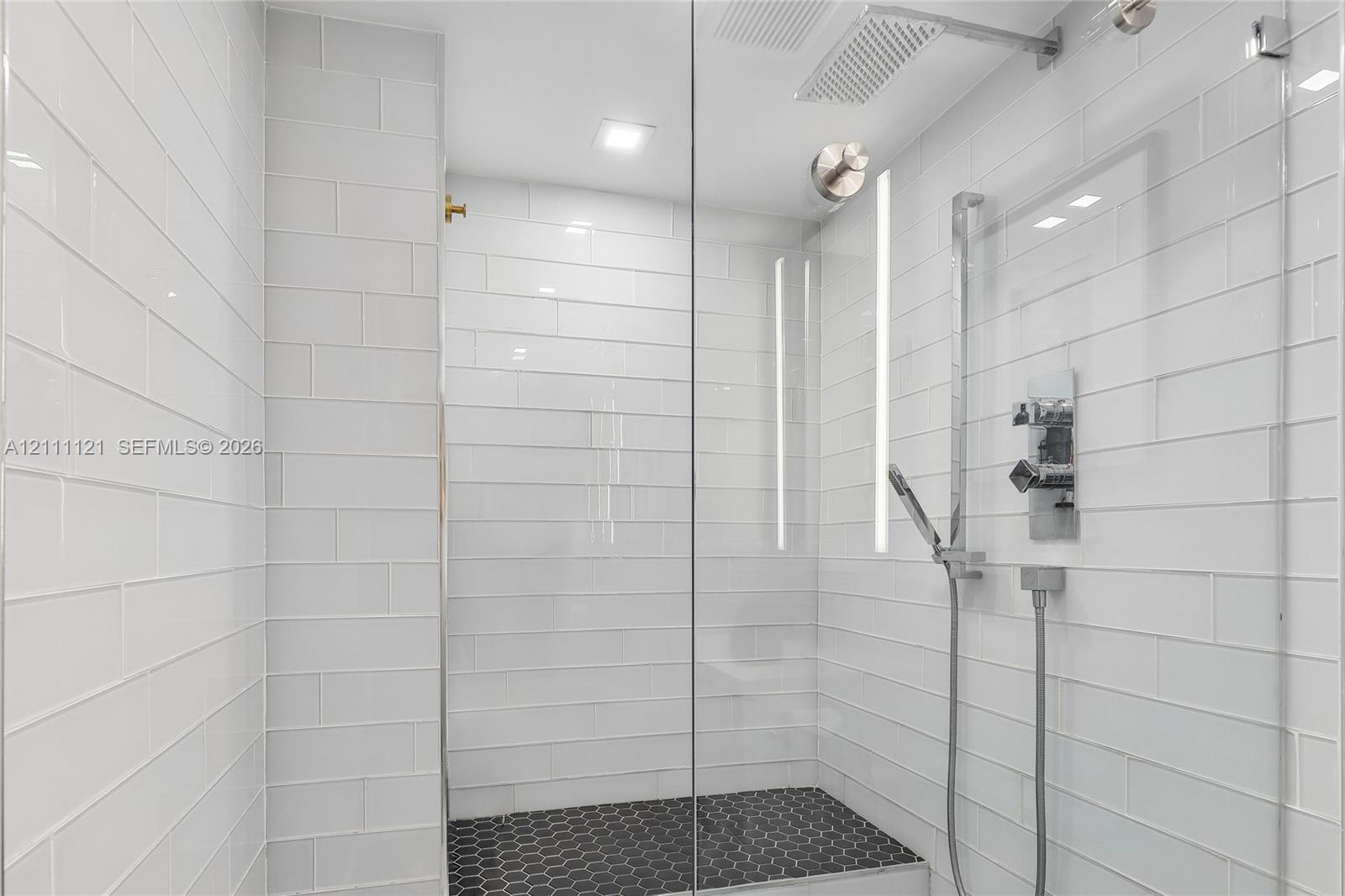 Walk-in shower featuring white subway wall tile, black hexagonal floor tile, a glass enclosure, a rainfall showerhead, and a handheld shower fixture