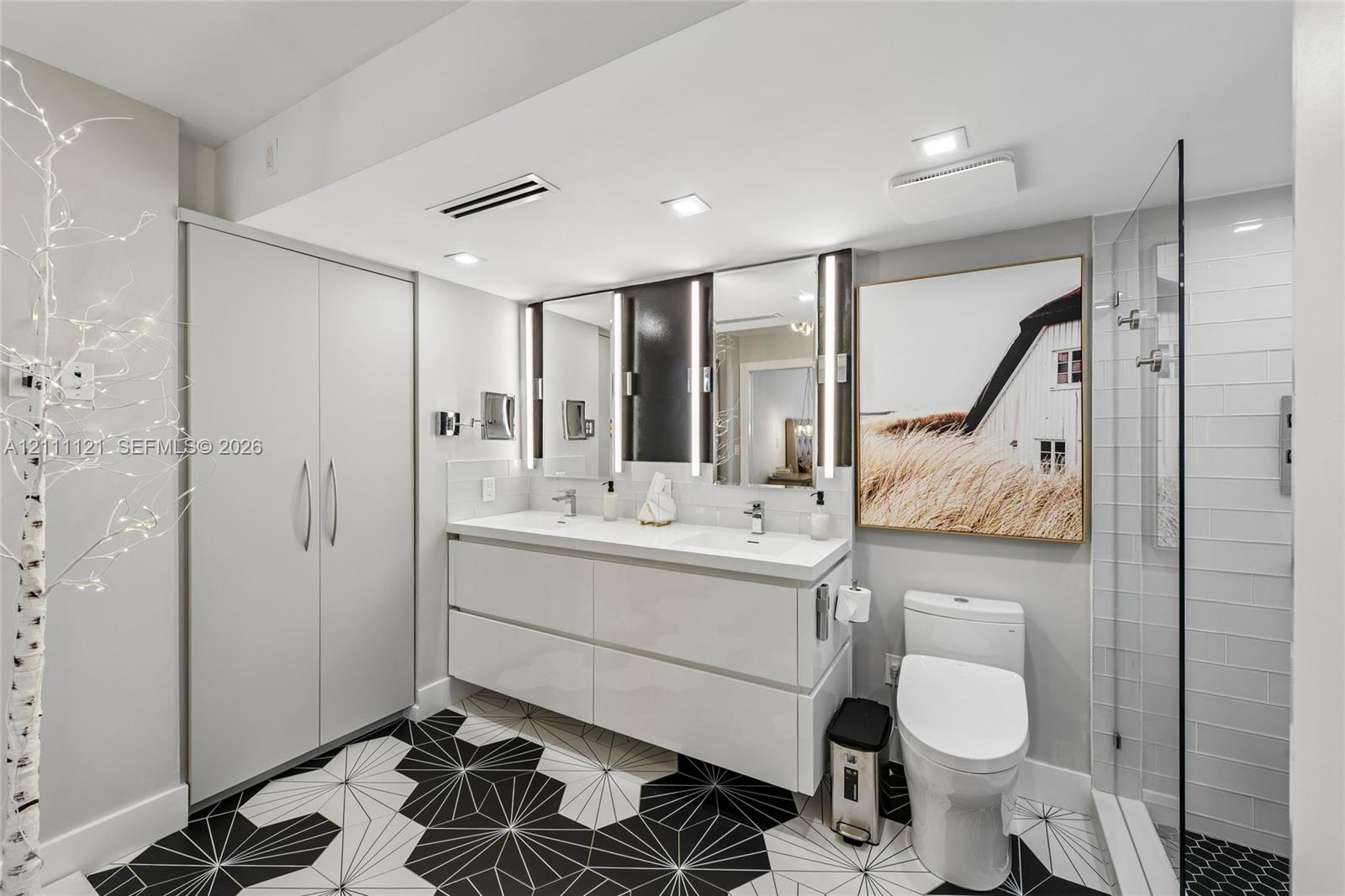 Floating double vanity with integrated sinks, recessed LED vanity lighting, floor-to-ceiling cabinetry, geometric tile flooring, and glass-enclosed shower with white subway tile surround
