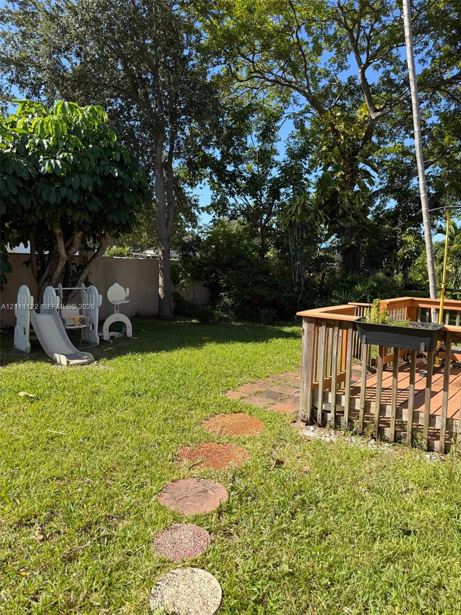 Private backyard featuring a wooden deck, stone-style pavers, a manicured lawn, and mature trees enclosed by a solid perimeter wall