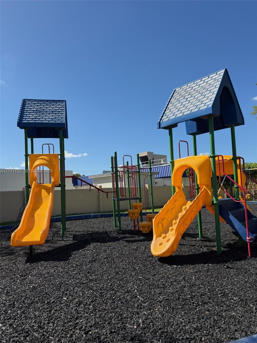 Outdoor recreation area featuring two multi-level play structures, molded slides, and ground-level mulch surfacing, enclosed by a perimeter wall