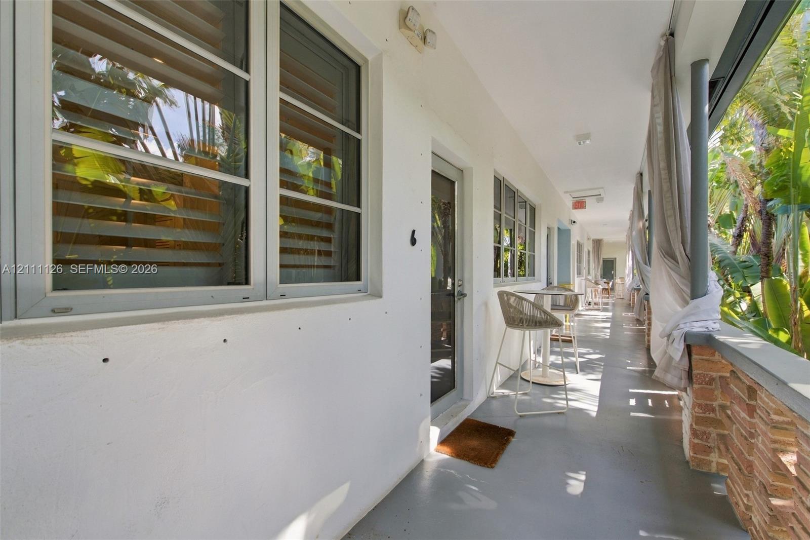 Covered exterior walkway featuring a smooth concrete floor, white stucco wall finish, metal-frame windows, and a brick-accented railing
