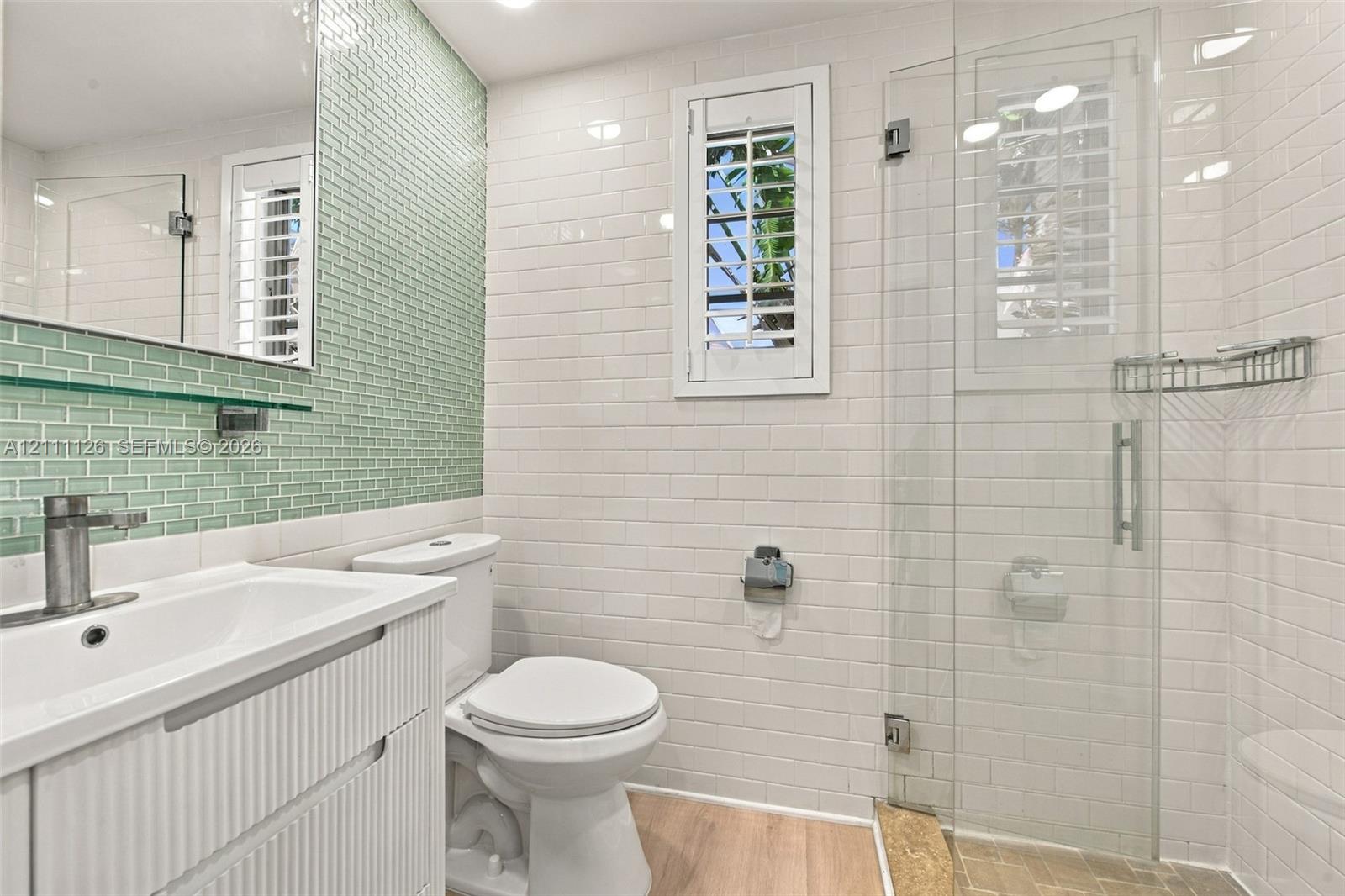 Bathroom featuring a vanity with integrated sink, light green glass mosaic wall tile, white subway tile shower surround, glass-enclosed shower, and wood-finish flooring