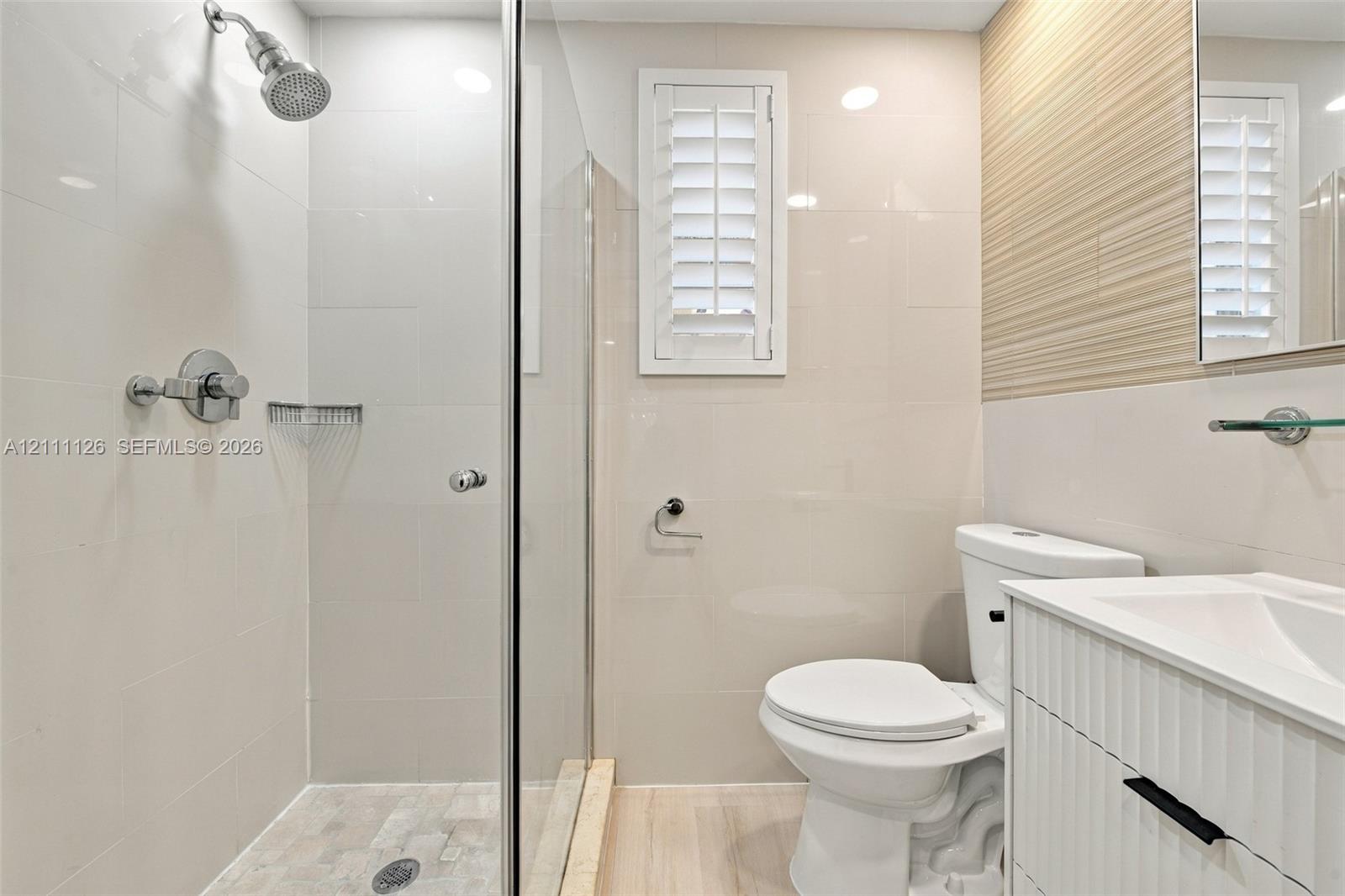 Walk-in shower with glass enclosure, wall-mounted shower head, textured accent wall, integrated vanity with white cabinetry, and shutter-style window treatment
