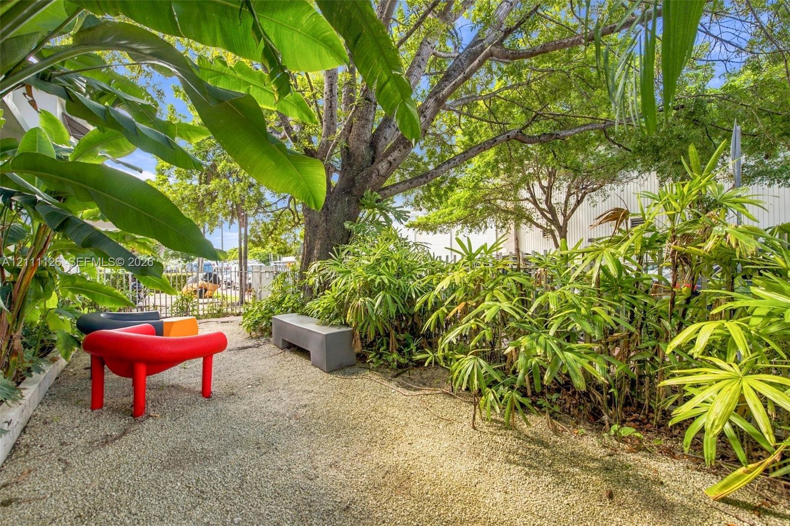 Private garden area featuring crushed gravel ground cover, a built-in stone bench, mature trees, and lush tropical foliage enclosed by metal perimeter fencing