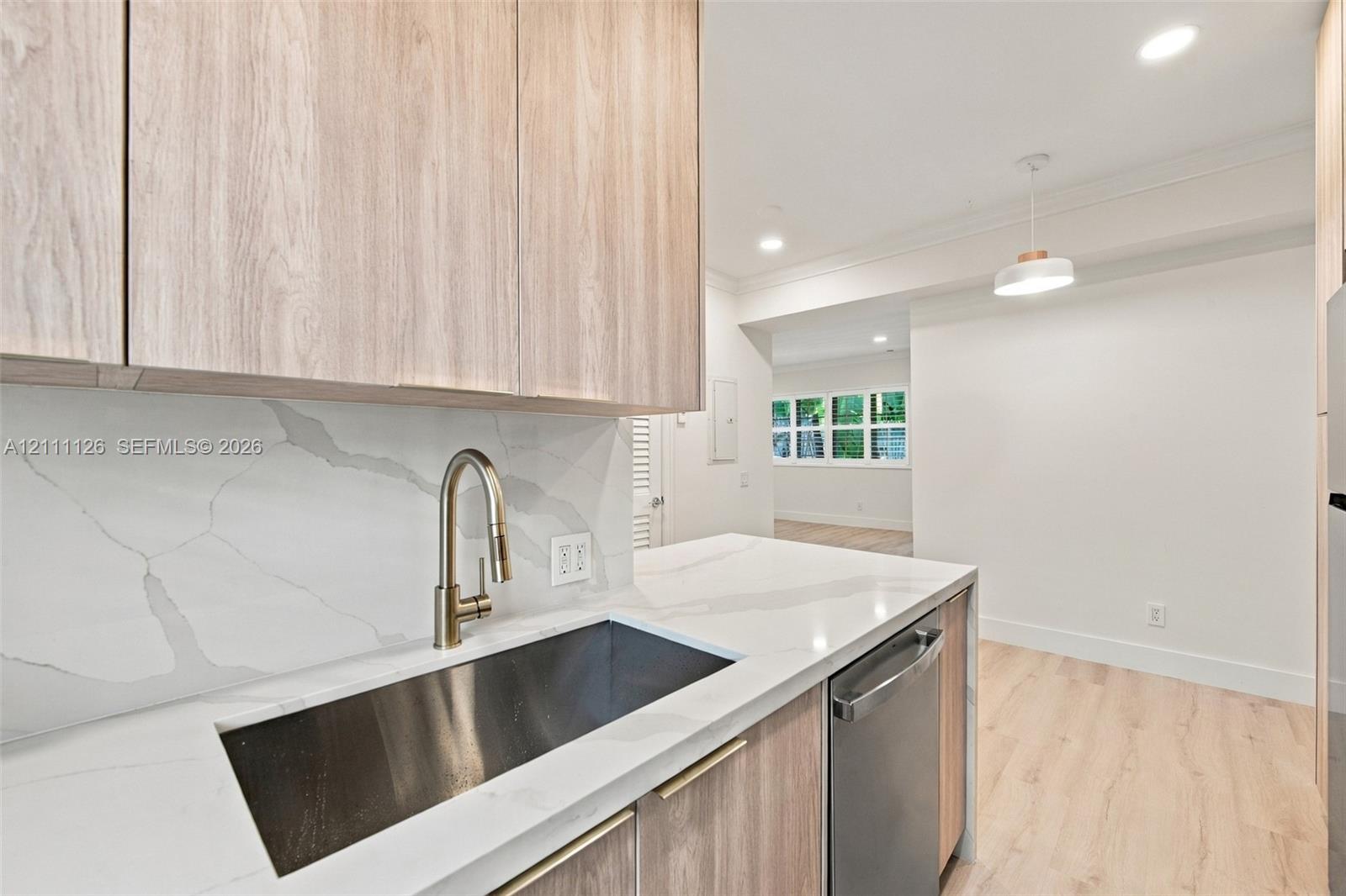 Kitchen featuring light wood-finish cabinetry, stone-surface countertops, full-height stone-surface backsplash, undermount stainless steel sink with a high-arc faucet, and recessed lighting