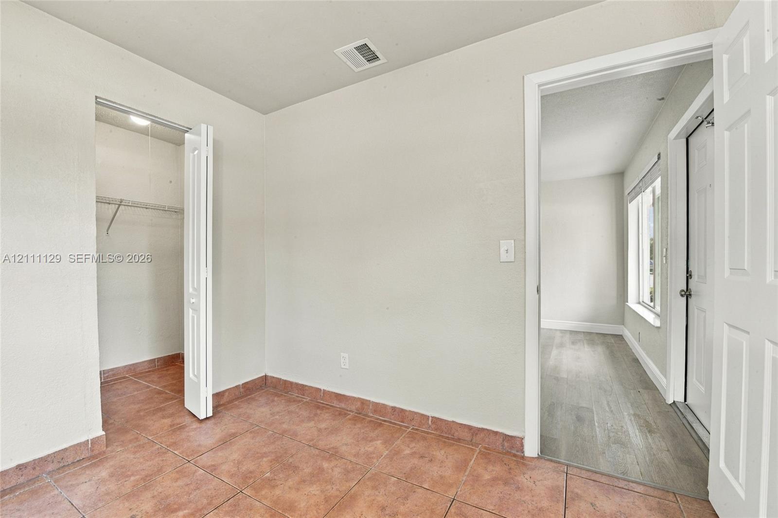 Interior room featuring terracotta-toned tile flooring, a walk-in closet with built-in shelving, and a doorway leading to a space with wood-finish flooring and a white paneled door