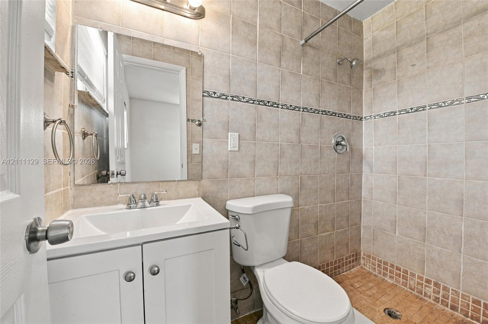 Bathroom featuring a vanity with an integrated sink, wall-mounted mirror, neutral tile wall surround with decorative accent inlay, and a built-in shower head