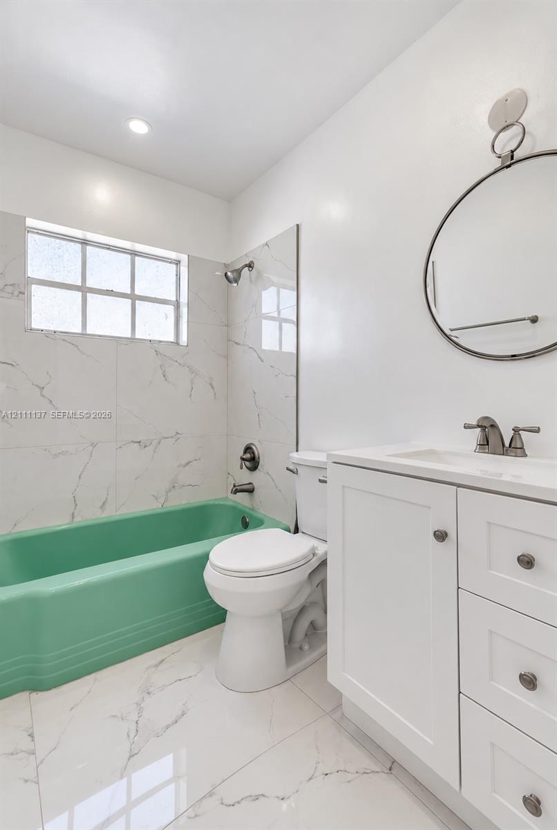 Bathroom featuring white tile flooring and wall surrounds with grey veining, a colored soaking tub, a white vanity cabinet with a stone-finish countertop, and a circular wall-mounted mirror