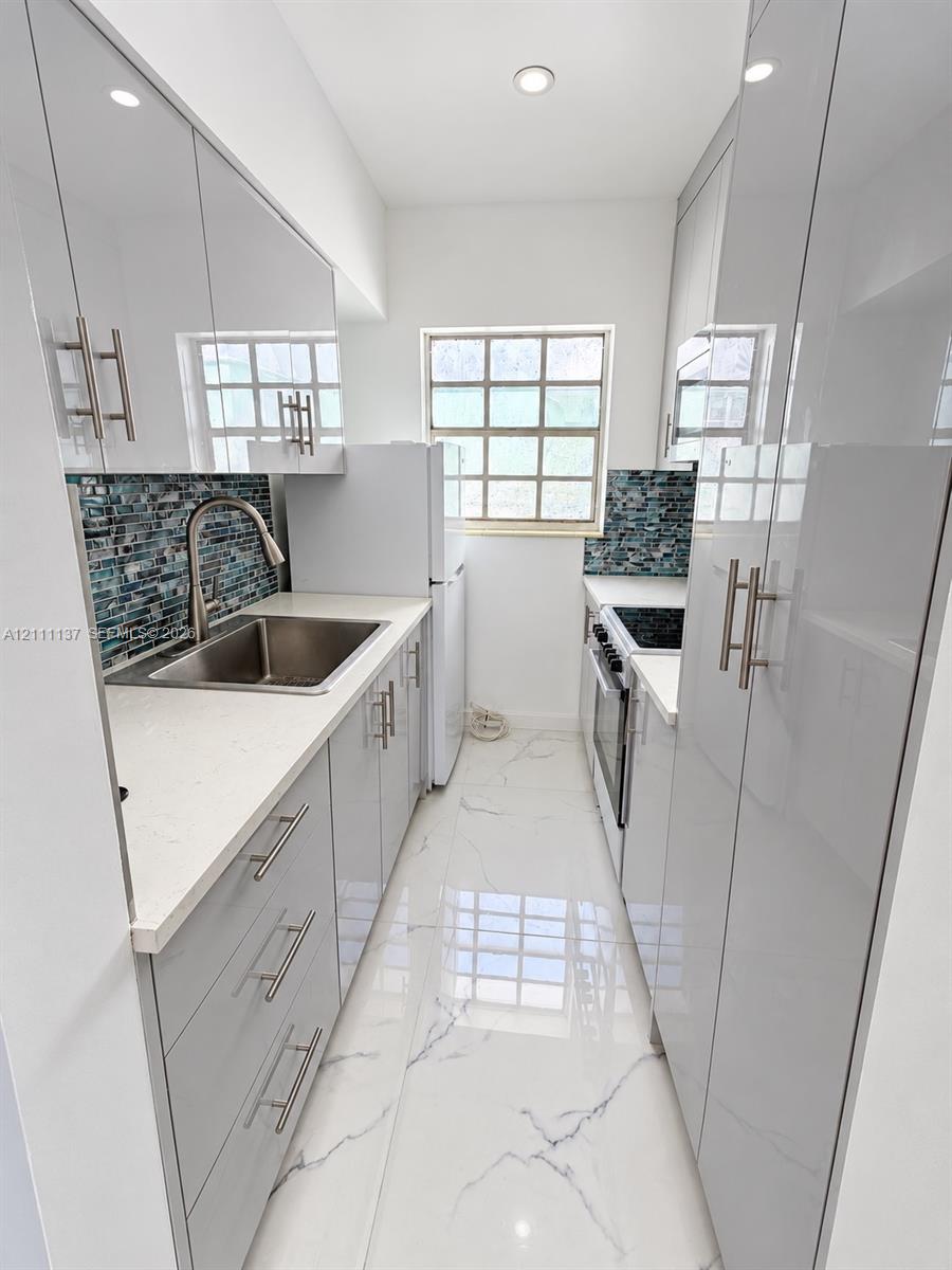Kitchen featuring high-gloss cabinetry, stone-surface countertops, blue mosaic tile backsplash, stainless steel sink with gooseneck faucet, and large-format porcelain tile flooring Kitchen featuring high-gloss cabinetry, stone-surface countertops, blue mosaic tile backsplash, stainless steel sink with gooseneck faucet, and large-format porcelain tile flooring