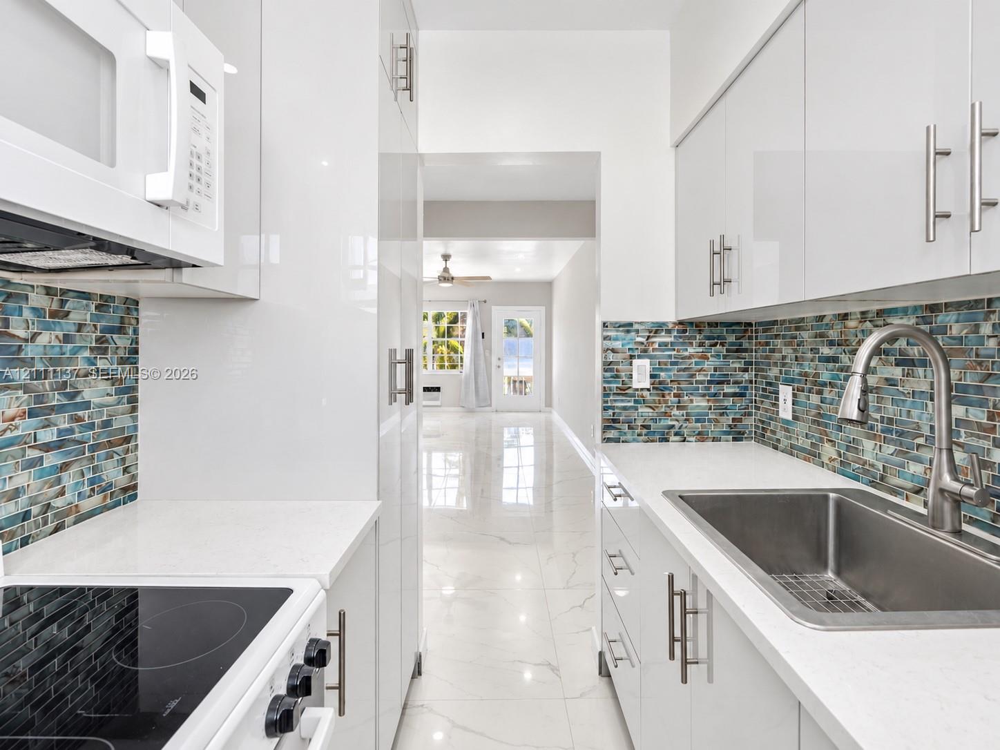 Kitchen featuring high-gloss white cabinetry, white quartz countertops, multi-tonal glass tile backsplash, stainless steel undermount sink with arched pull-down faucet, and polished stone-finish flooring Kitchen featuring high-gloss white cabinetry, white quartz countertops, multi-tonal glass tile backsplash, stainless steel undermount sink with arched pull-down faucet, and polished stone-finish flooring