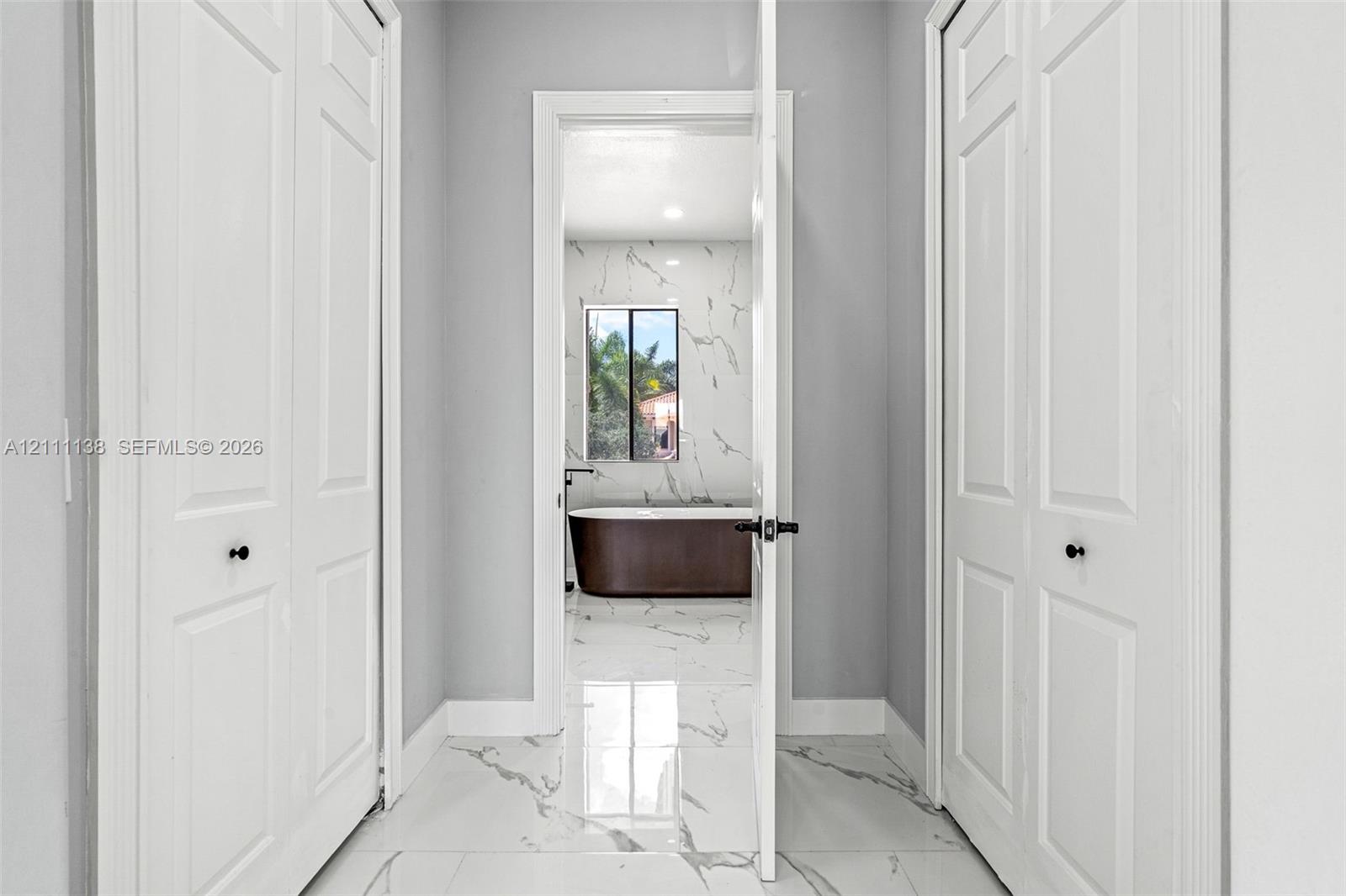 Hallway featuring white panel doors with matte black hardware and polished stone-finish floor tile with veining, leading into a space with matching stone-finish wall tile and a freestanding soaking tub